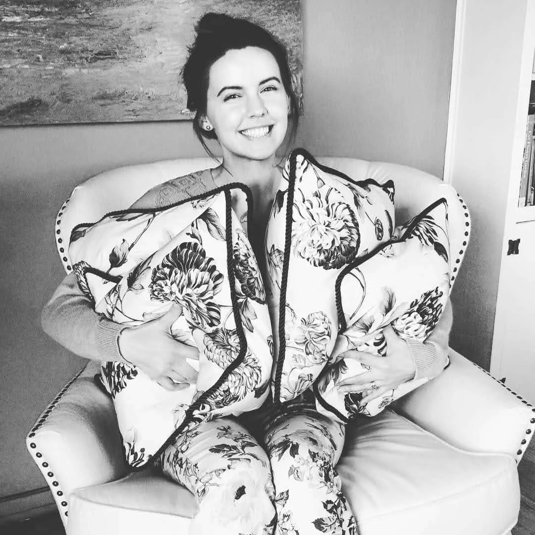 A smiling woman named Chelsea Labanowski with dark hair sitting on an upholstered chair hugging a pile of handmade pillows with art in the background.