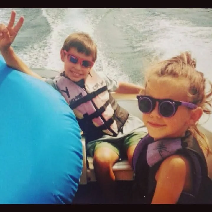 Two children, wearing sunglasses and life jackets, are on a boat ride on the water, smiling and waving.