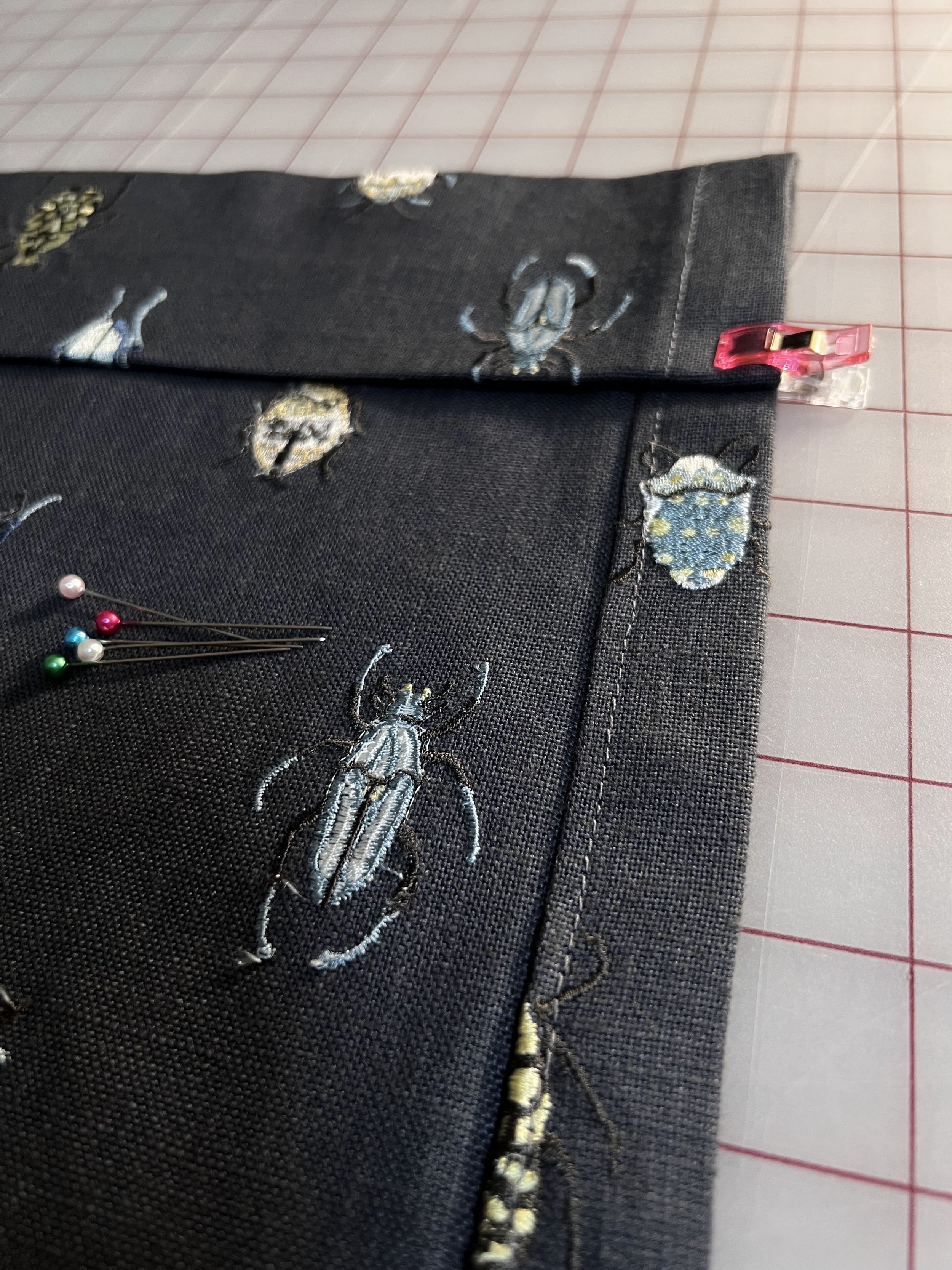 Detail shot of dark blue fabric with an embroidered bug design, fabric clip holding fabric, pin with colored heads on fabric, and cutting mat in background.