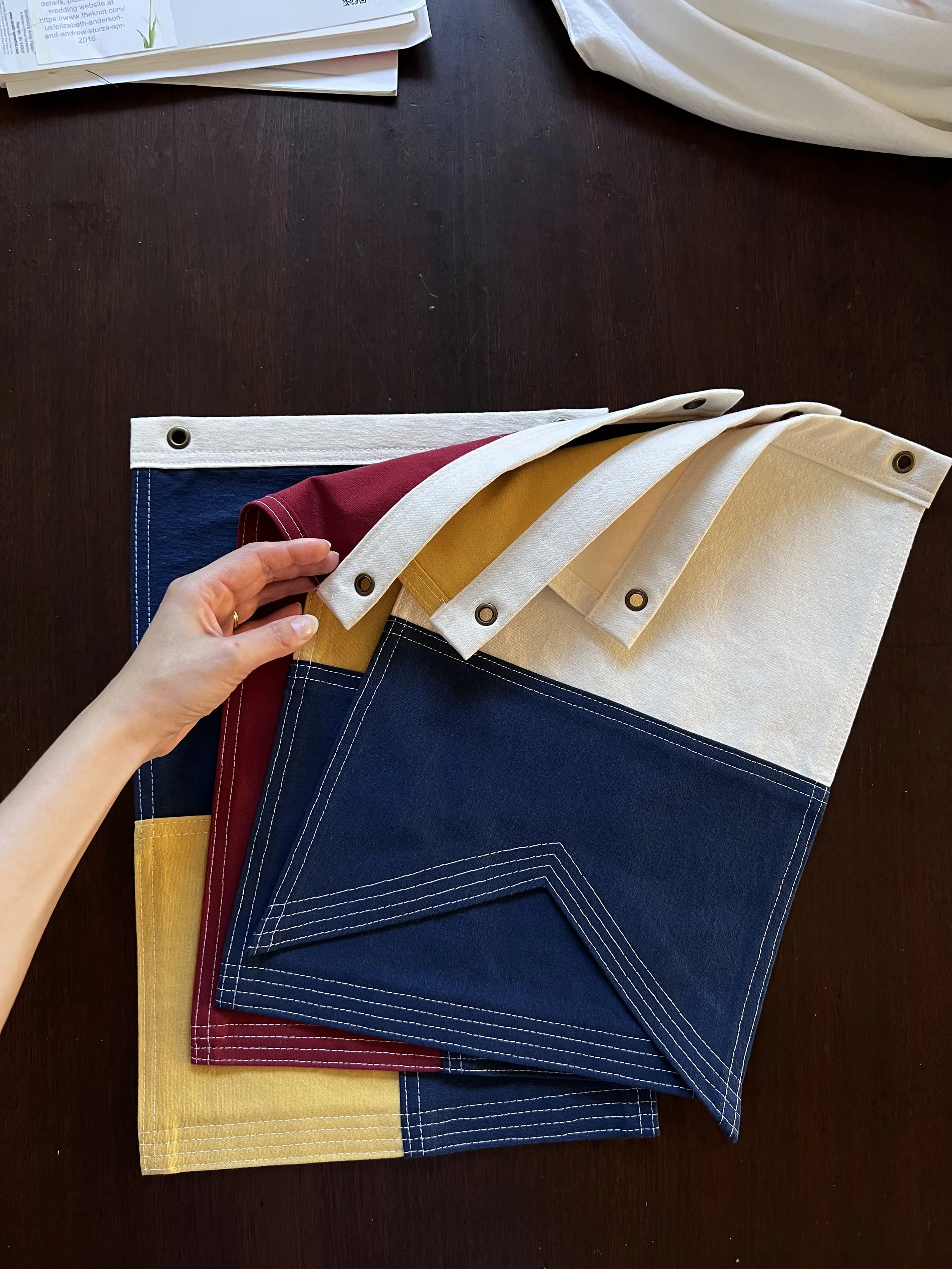 A hand thumbing through a stack of nautical flags "L", "A", "K", and "E" in blue, red, yellow, and off-white, each with grommets and white stitching, on a dark wooden surface.