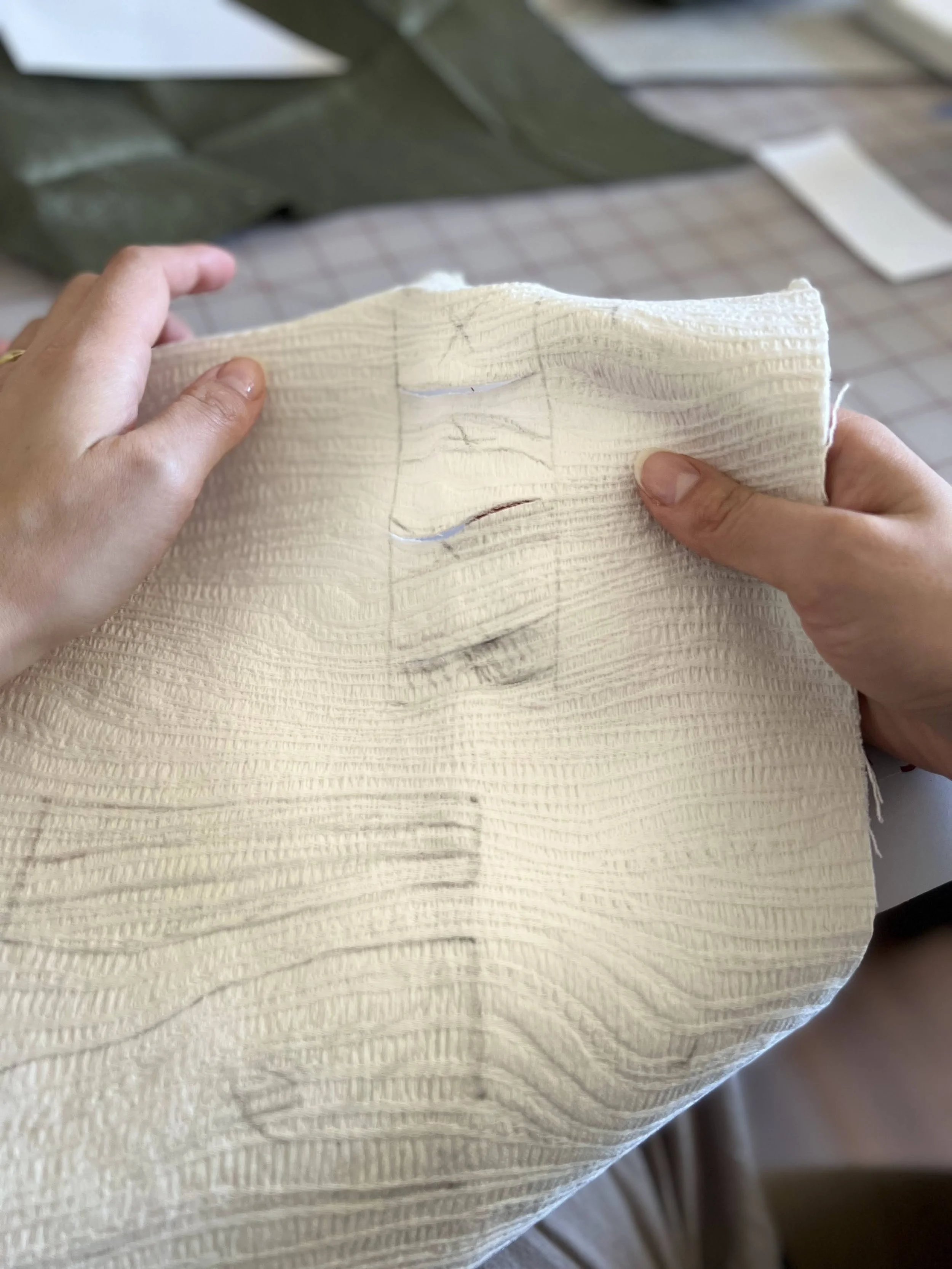 Close-up of a work-in-progress sewing project with a craftsperson's hands holding a textured, cream-colored fabric with pencil markings.
