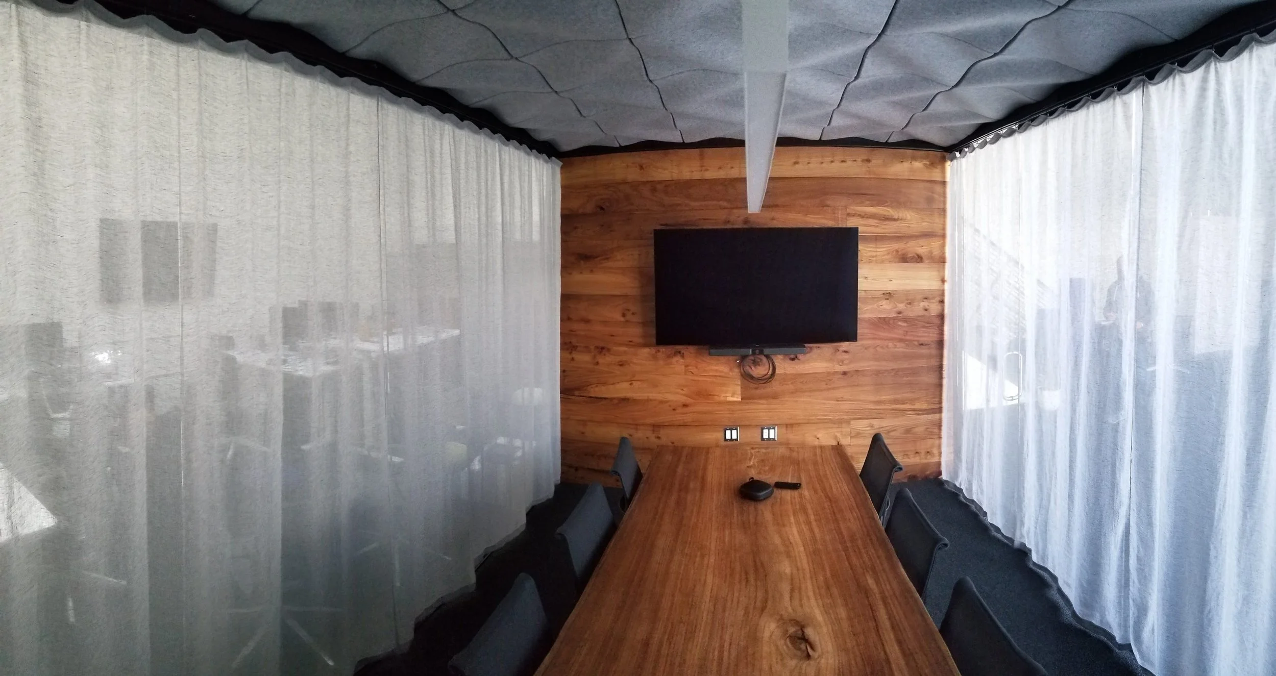 A meeting room with a long wooden table surrounded by chairs, a wall-mounted television, and custom sheer curtains covering large windows, with wood paneling on the wall behind the TV and acoustic panels on the ceiling.