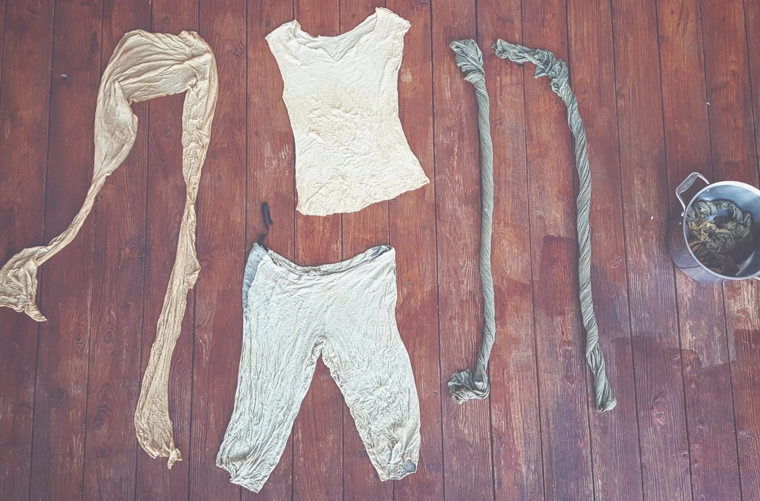 Handmade costume for Ray from Star Wars laid out on a wooden floor, including a distressed beige shirt, distressed gray pants, distressed gray arm wraps, a scarf from Jakku, with a pot for hand-dyeing natural fabric.