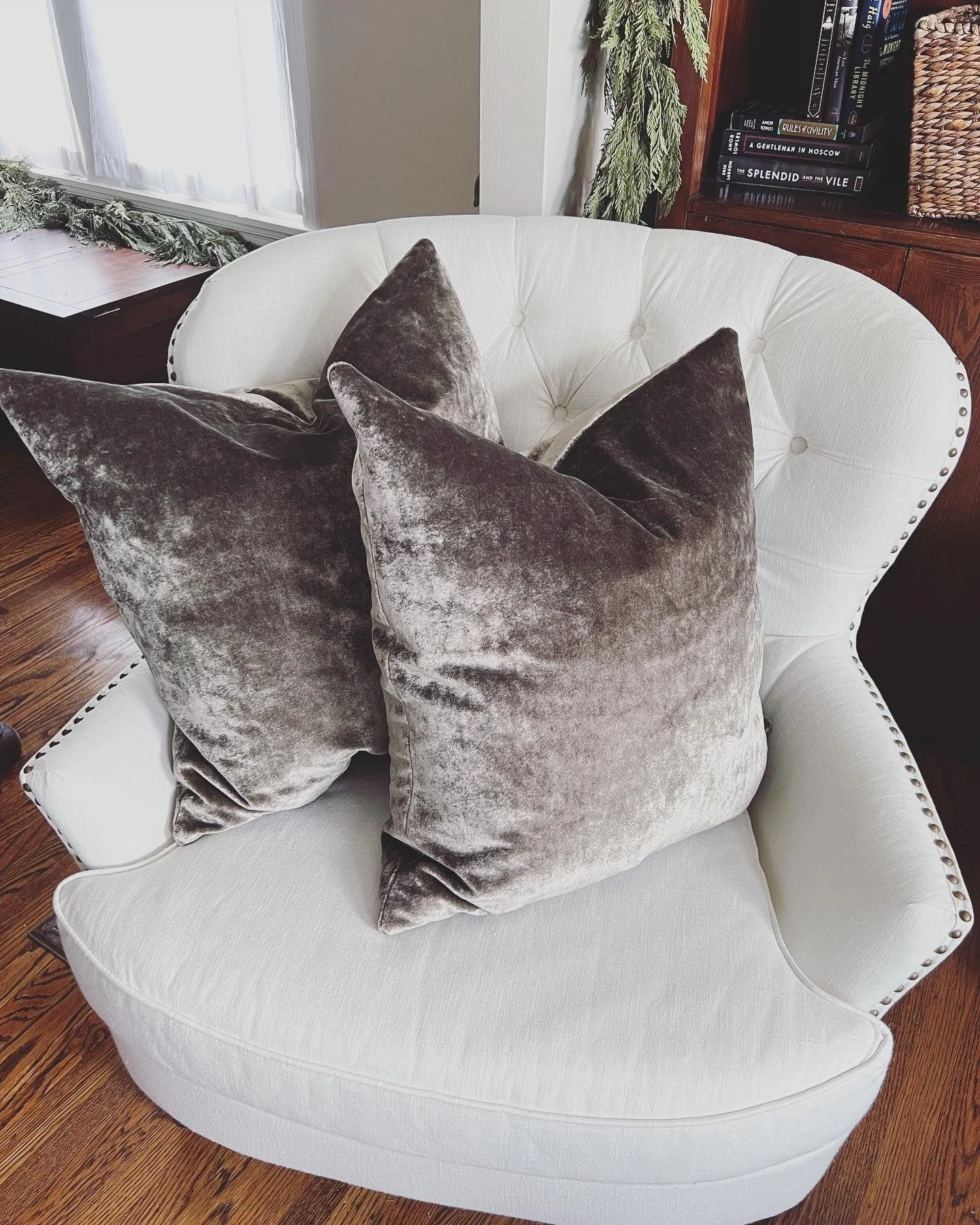 White upholstered Pottery Barn armchair with tufted back and studded trim, adorned with two velvet gray pillows, situated on a wooden floor in front of a bookcase near a window with handmade white cafe curtains.