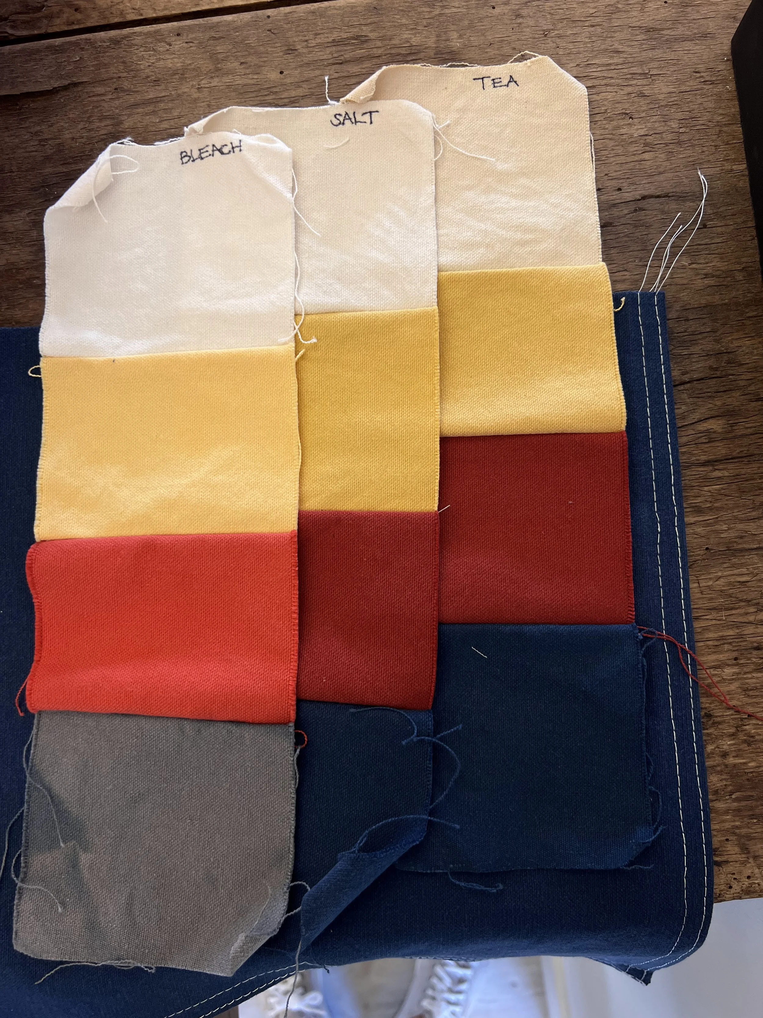 Distressed swatches of nautical flag fabric labeled with varying methods of distress: bleach, salt, and tea. The swatches are in various colors, including off-white, yellow, red, and navy blue, on a wooden surface.