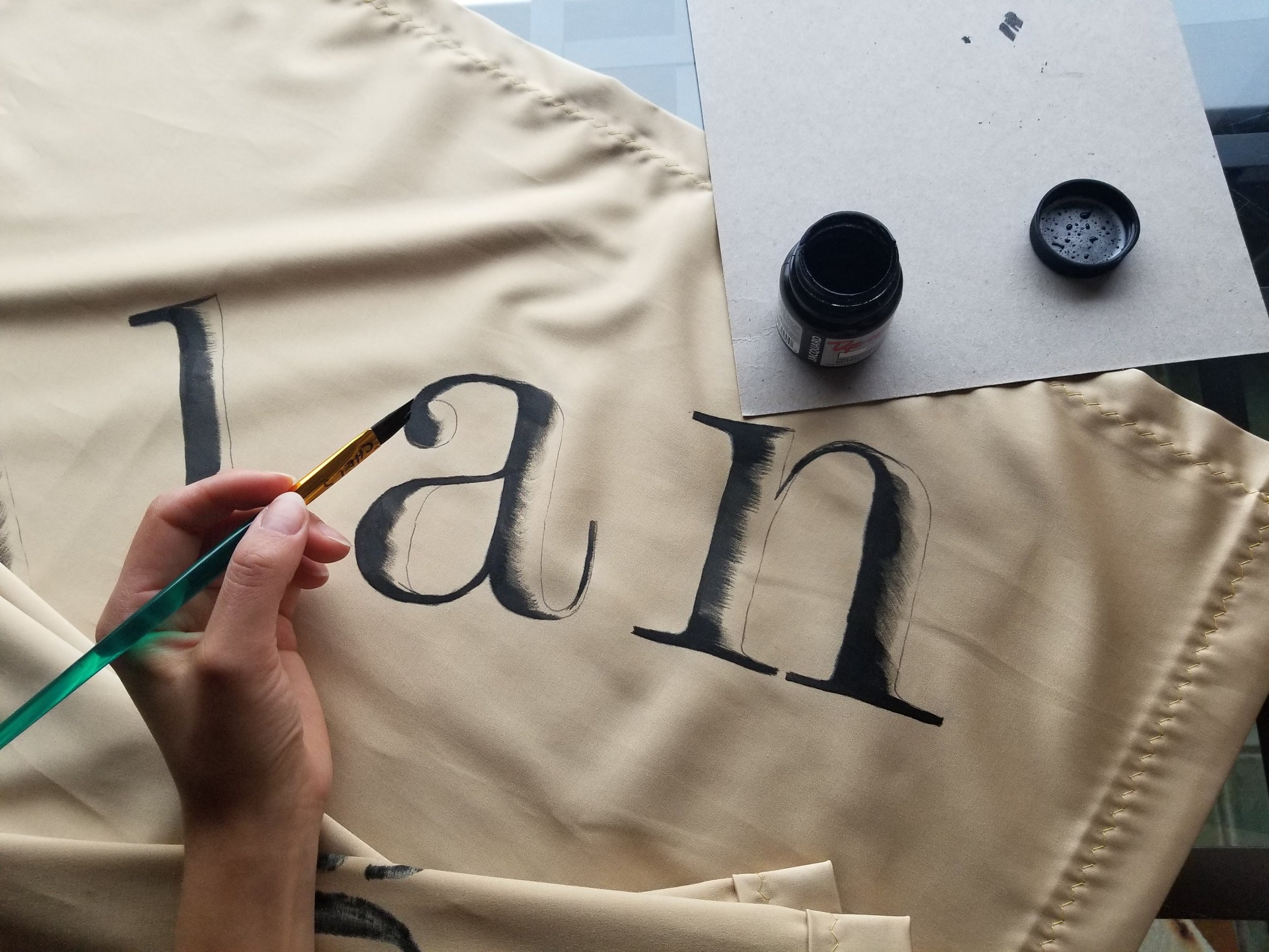 A left-handed artist is using a fine paintbrush to paint vintage black letters "an" on a gold fabric with India Ink. An open small black paint container and its lid are on a sheet of paper on the fabric.