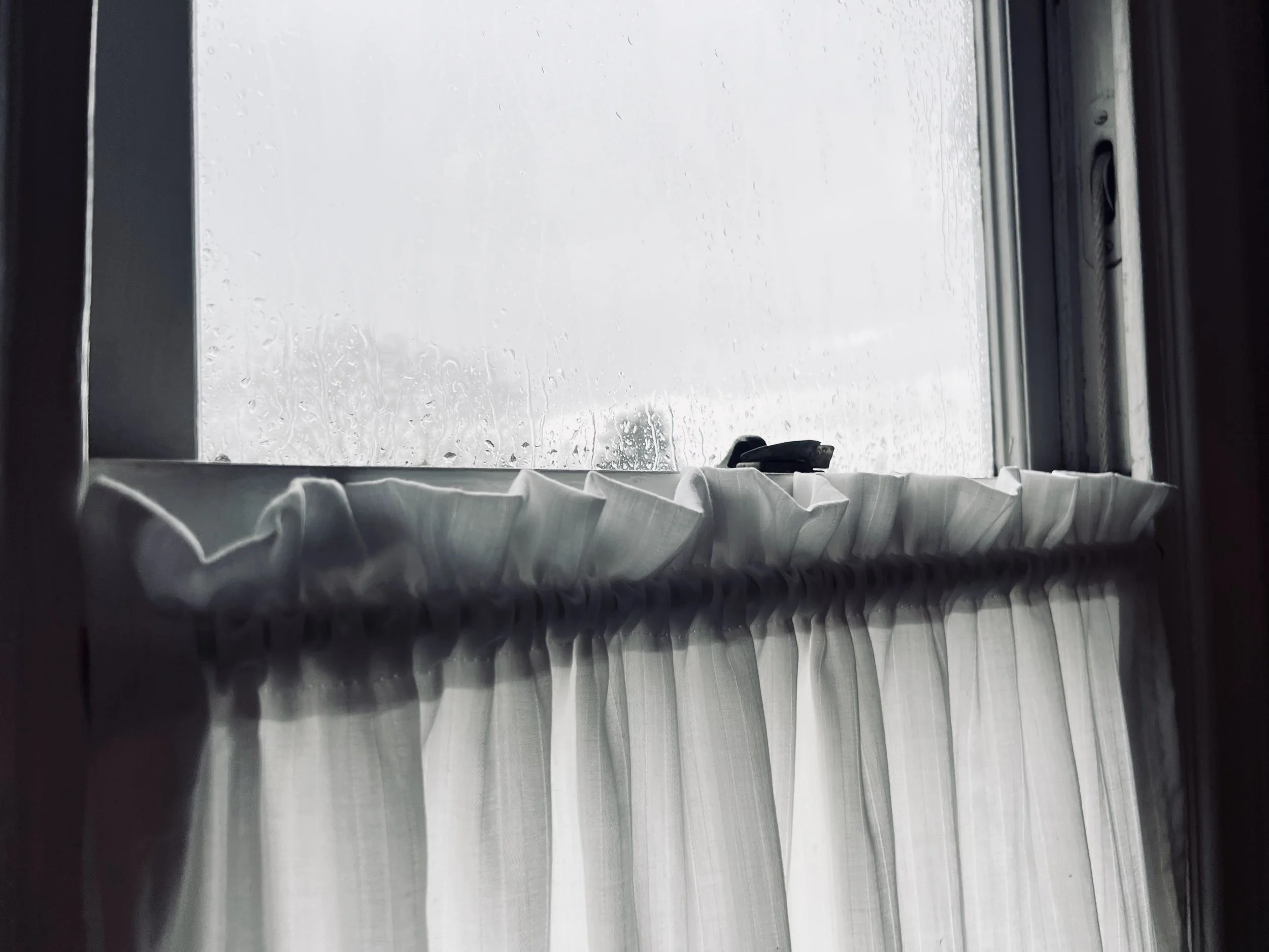 A moody view of rain streaking down a window on a rainy day, with blurred clouds and trees in the background.
