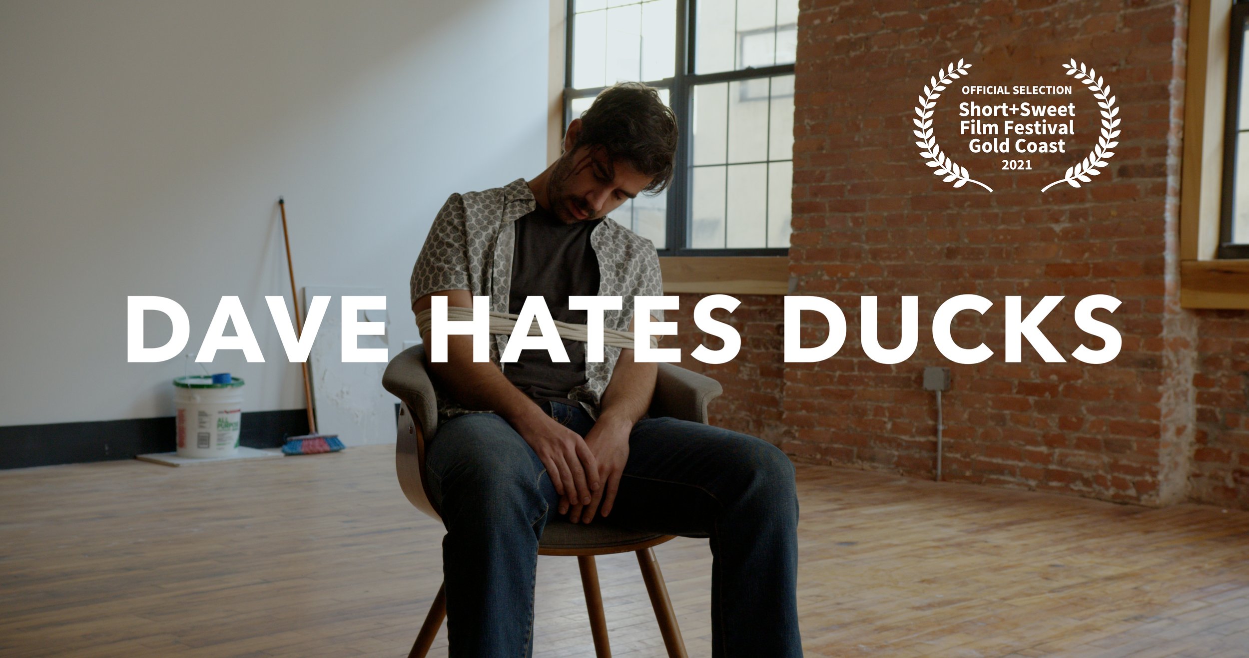 A young man with dark hair and a beard is sitting in a modern room with wooden floors, a brick wall, and large windows. He appears to be sad, with his head bowed down and hand resting on his lap. There is a large white text overlay that reads "DAVE H