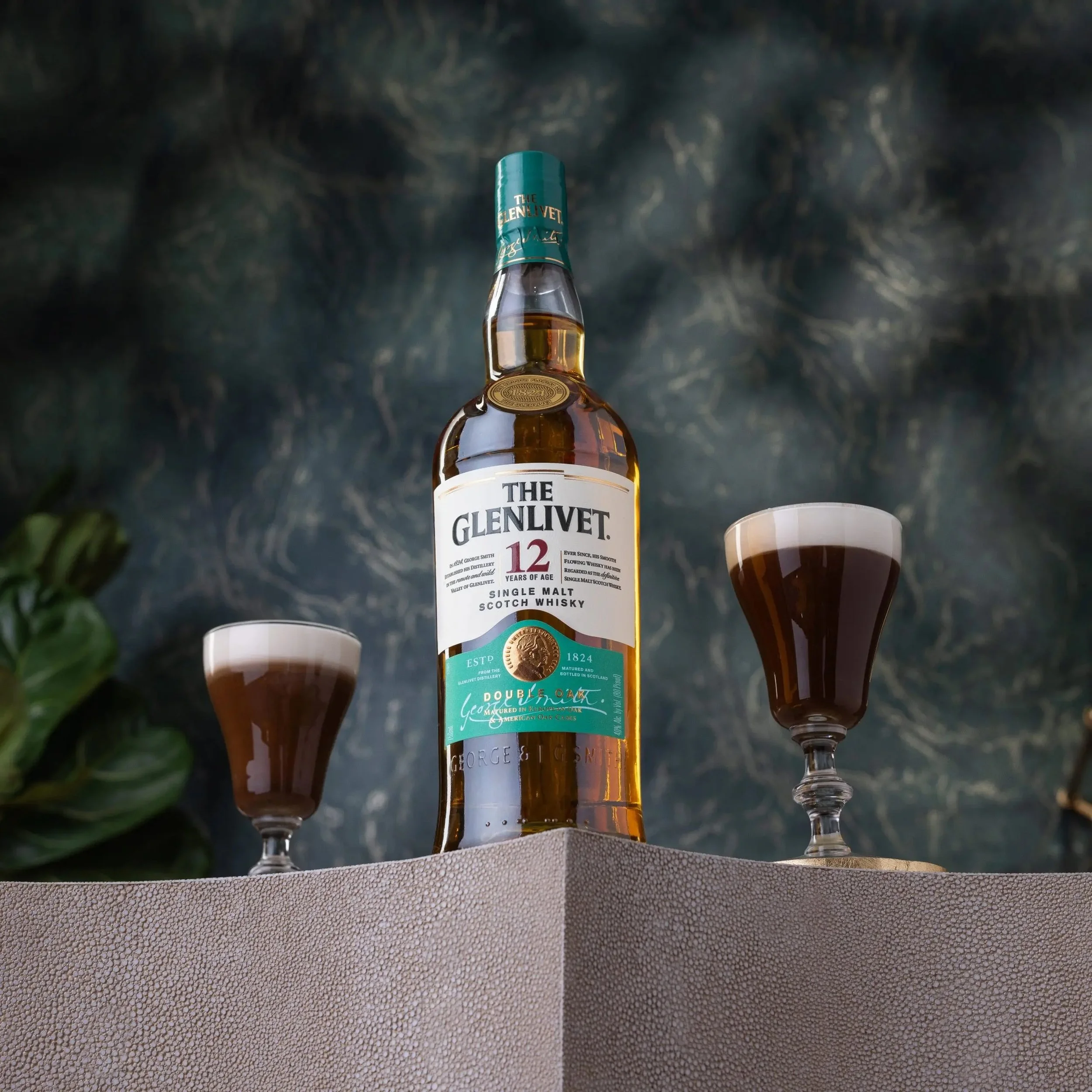 A bottle of The Glenlivet 12-year-old single malt Scotch whisky with two glasses filled with dark whisky, placed on a textured surface against a dark, marbled background.
