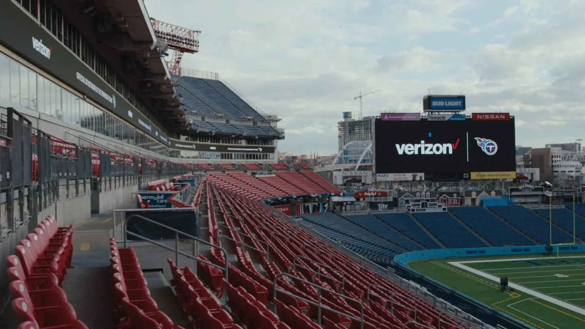 Empty football stadium with red and blue seats, a large digital scoreboard displaying Verizon and Tennessee Titans logos, and a cityscape in the background.