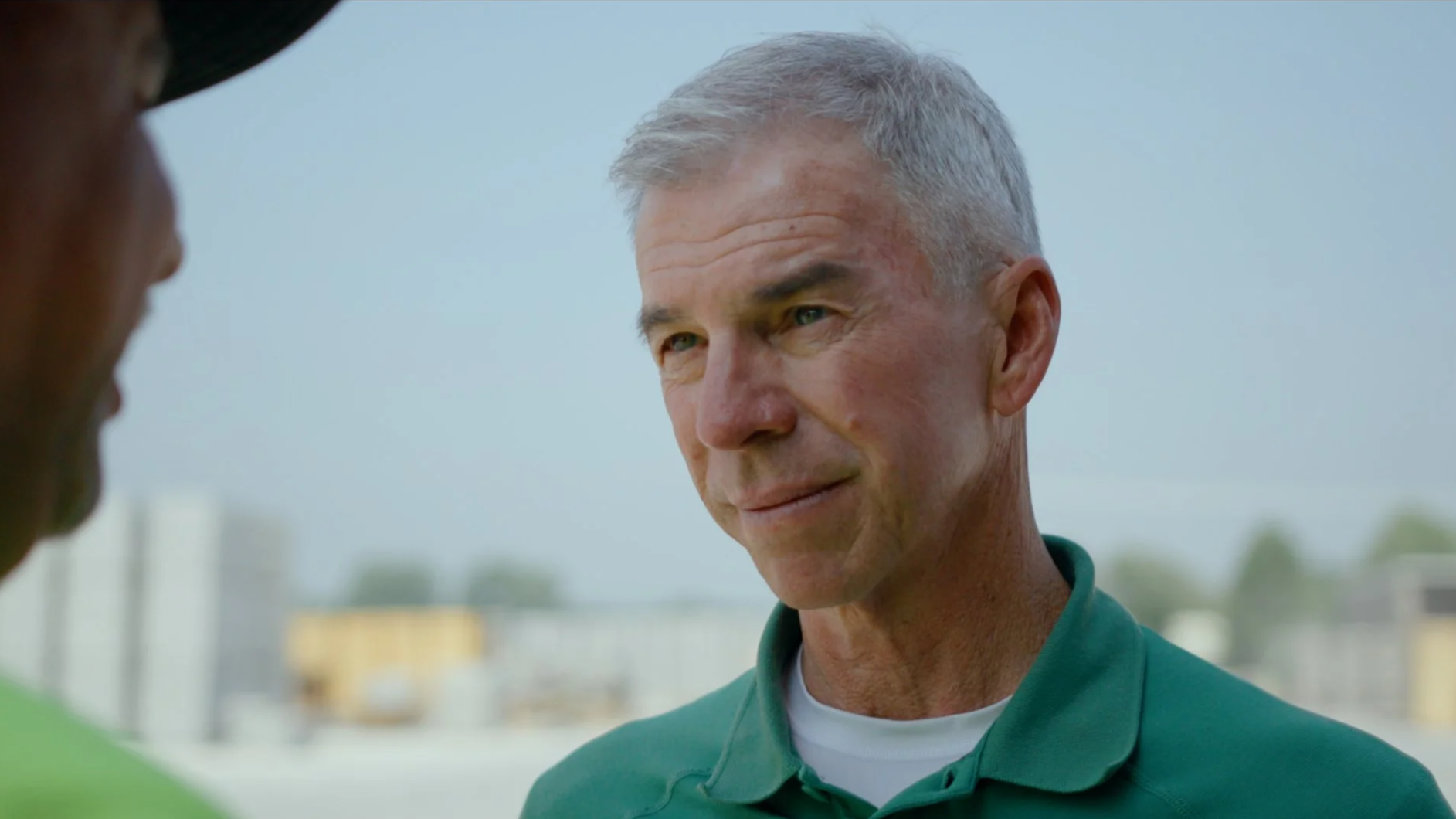 Close-up of an older man with short gray hair wearing a green collared shirt, looking at a person partially visible on the left side of the image, outdoors with a blurry cityscape background.