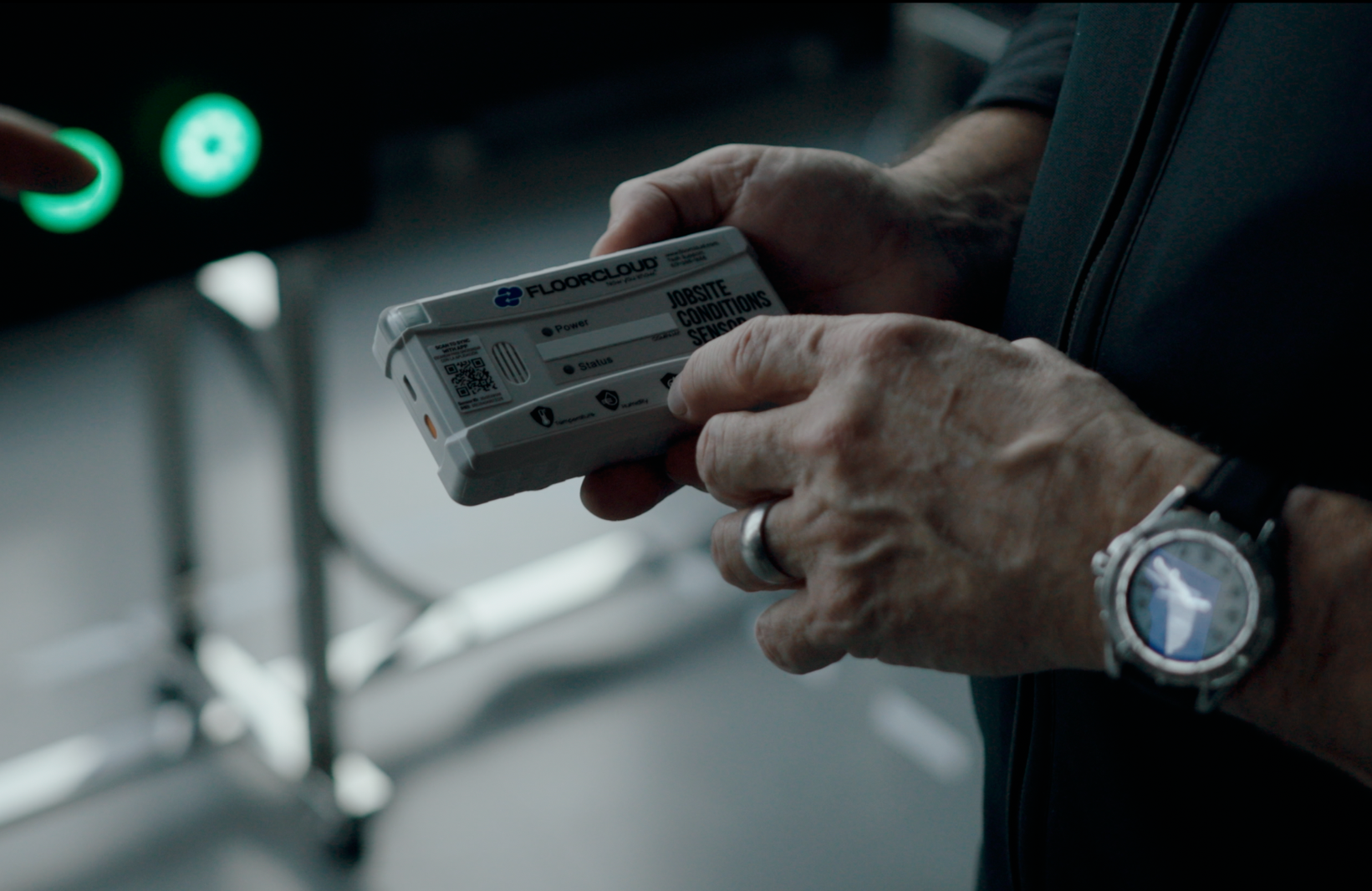 Close-up of a person holding a white device labeled 'FLOORCLOUD' with various icons and text, including 'JOBSITE CONDITIONS SENSOR'. The person is wearing a wristwatch and a ring.