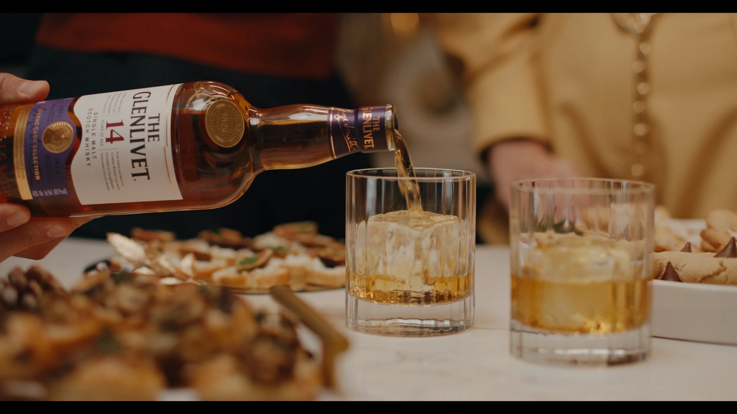 Person pouring The Glenlivet 14 scotch whisky into a glass with ice, on a table with snacks and desserts in the background.