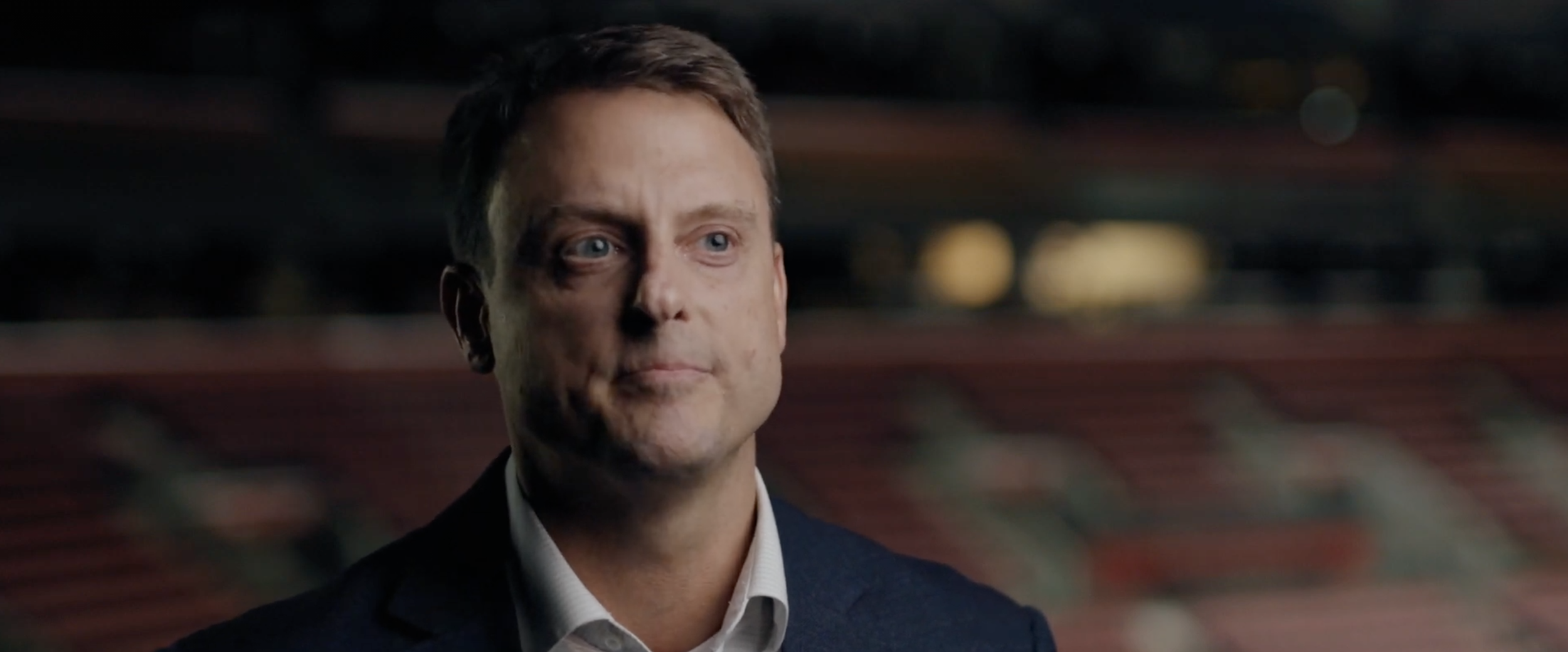 Close-up of a man with short brown hair, blue eyes, and wearing a dark suit jacket and white shirt, standing in a stadium with blurred seats in the background.