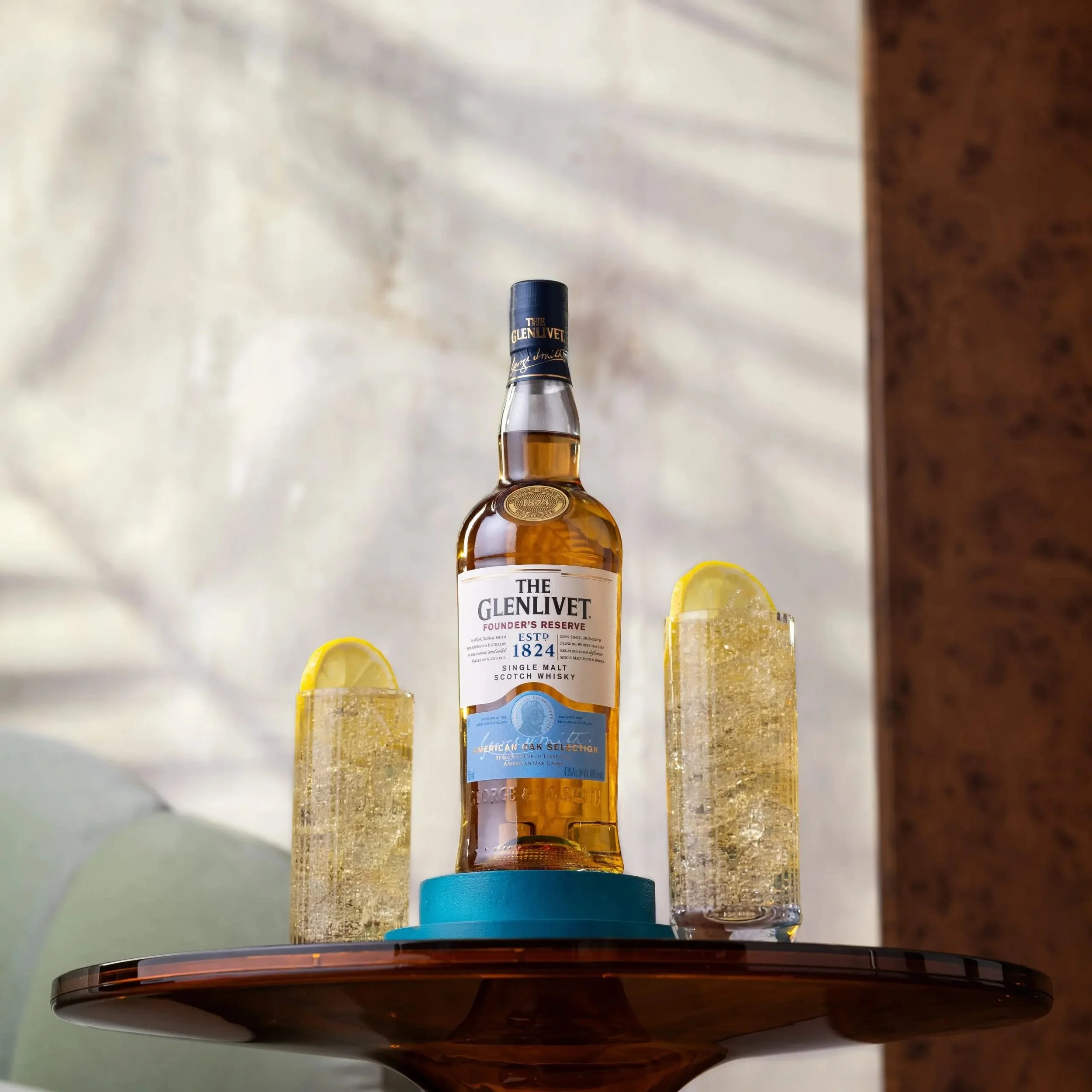 A bottle of The Glenlivet Founder’s Reserve Scotch Whisky on a small blue coaster, flanked by two glasses of sparkling drink with lemon slices, all on a round wooden table.