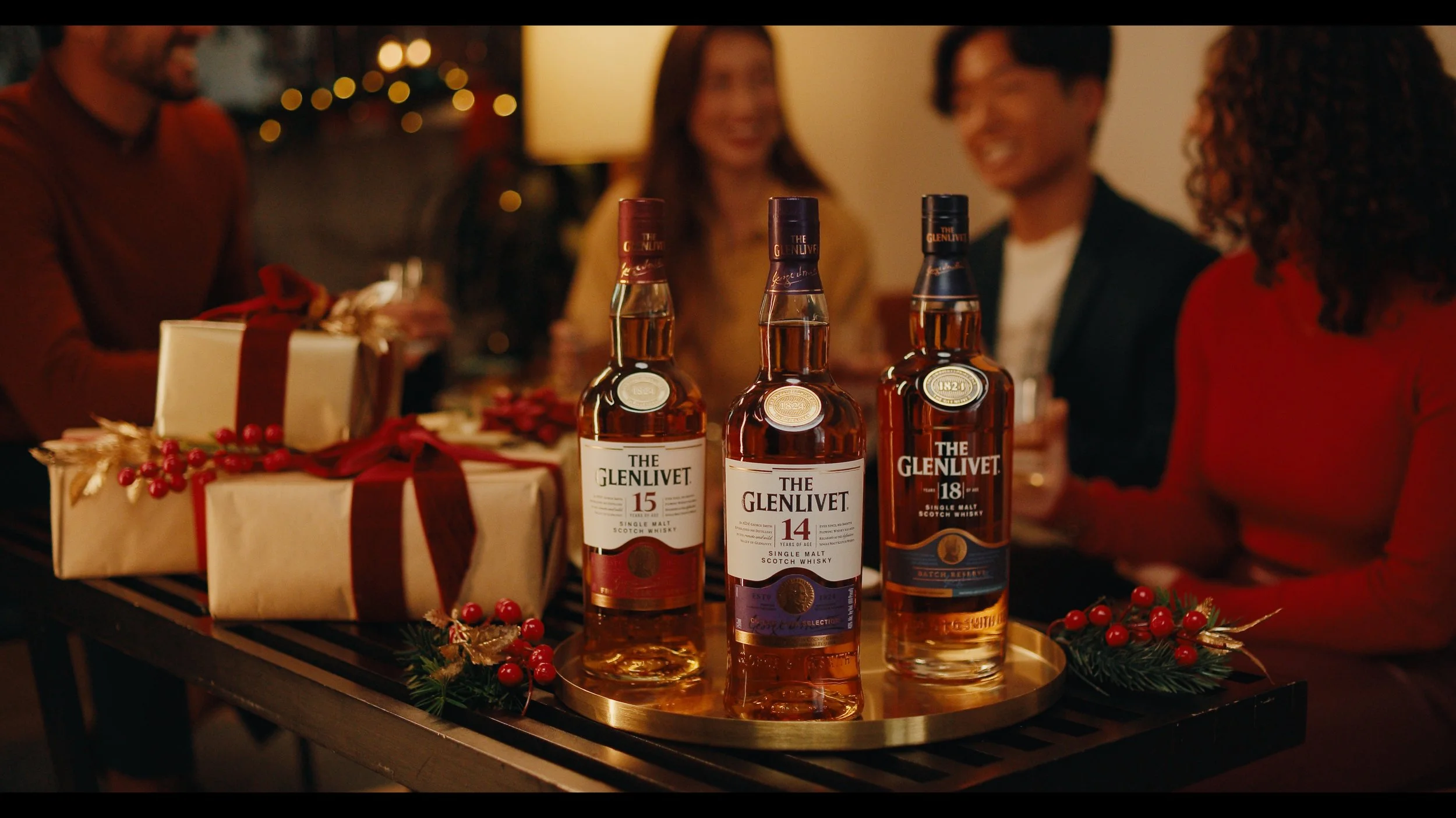 Three bottles of The Glenlivet Scotch whisky on a table surrounded by Christmas decorations and presents with four people in the background celebrating.