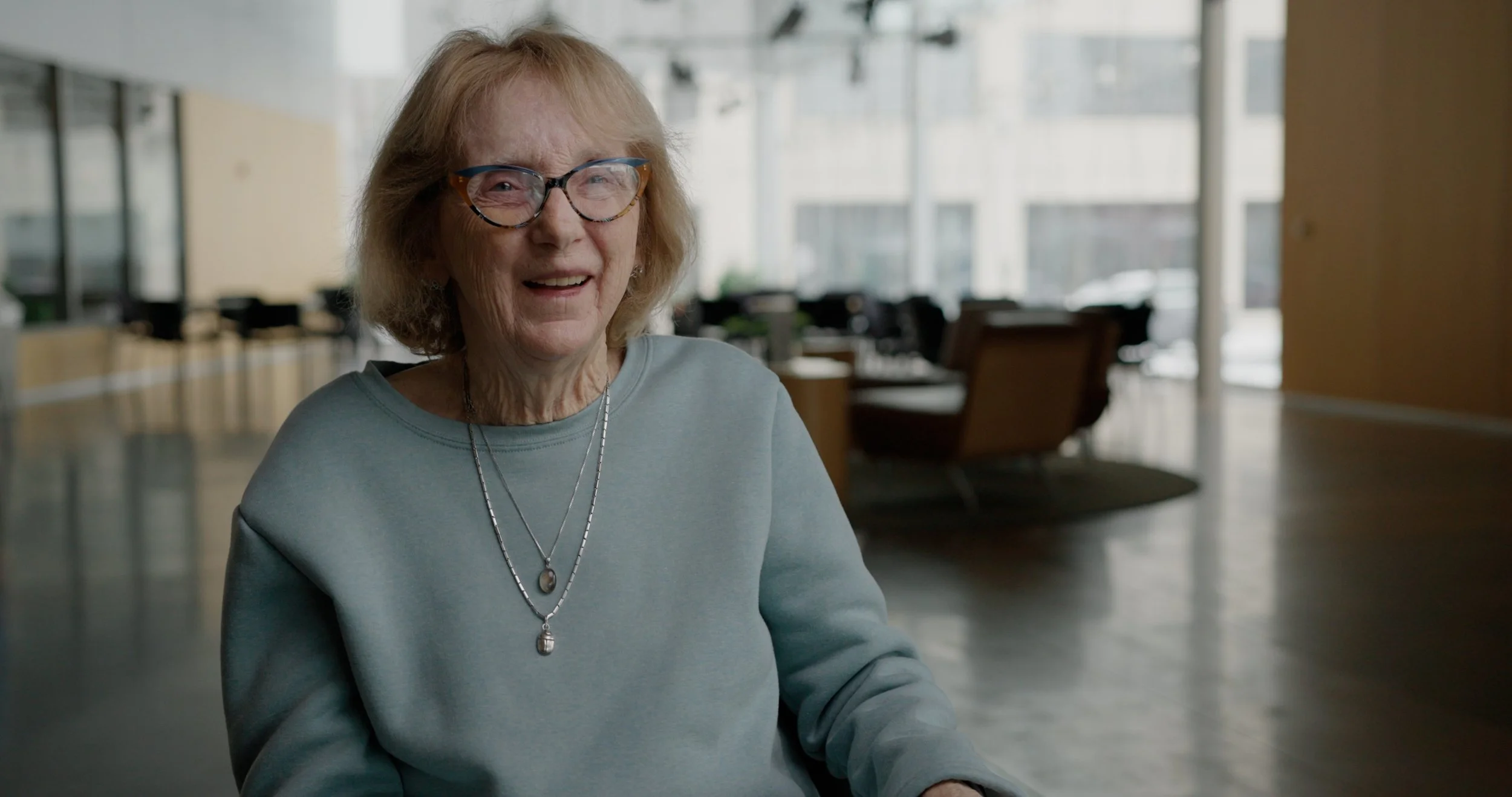 An elderly woman with glasses smiling, sitting indoors in a modern, well-lit space with large windows and comfortable chairs in the background.