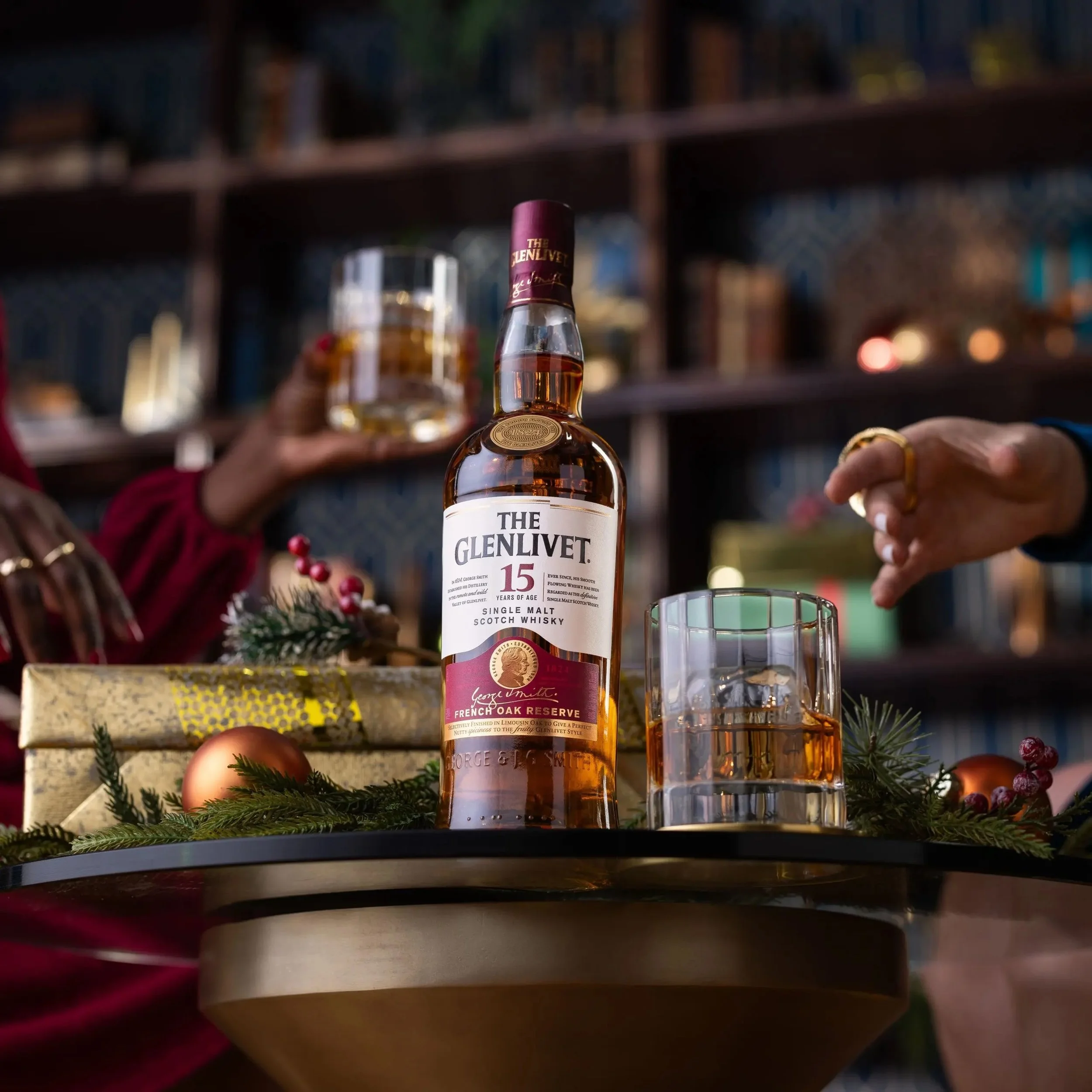 A bottle of The Glenlivet 15 Years Old Single Malt Scotch Whisky on a table, with two glasses of whiskey, holiday decorations, and hands holding a glass in the background.