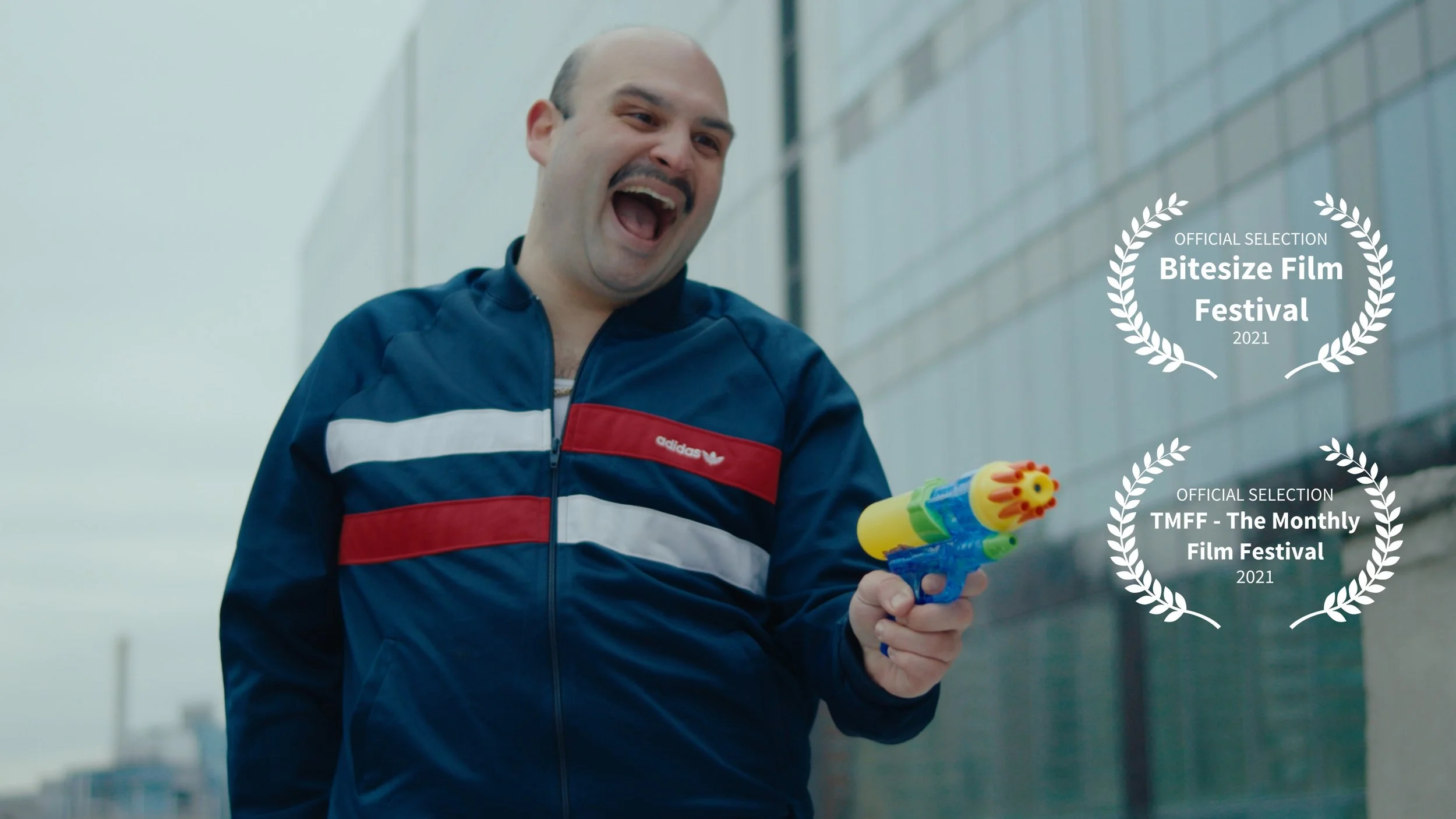 A man smiling and aiming a toy water gun, wearing a blue Adidas jacket, standing outdoors near a brick building.