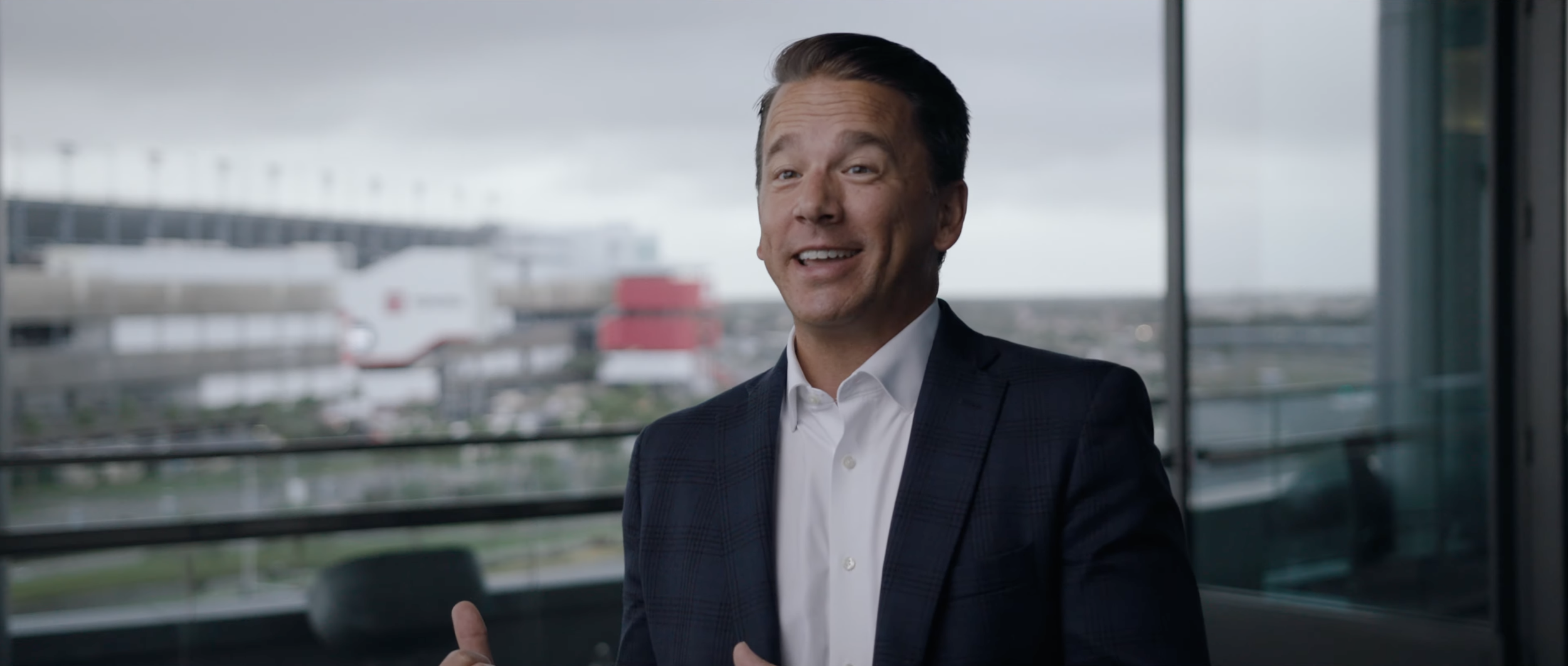 A man in a business suit smiling and speaking in an office with large windows and a city view.