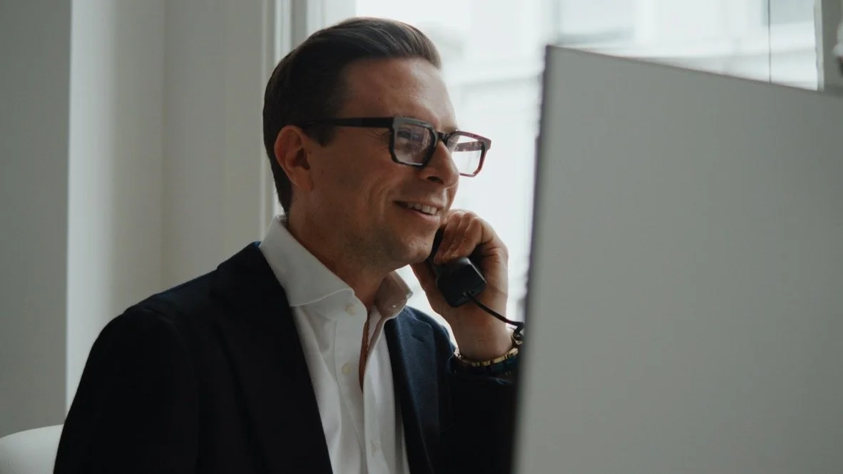 A man with dark hair, glasses, wearing a white shirt and dark blazer, smiling while talking on the phone