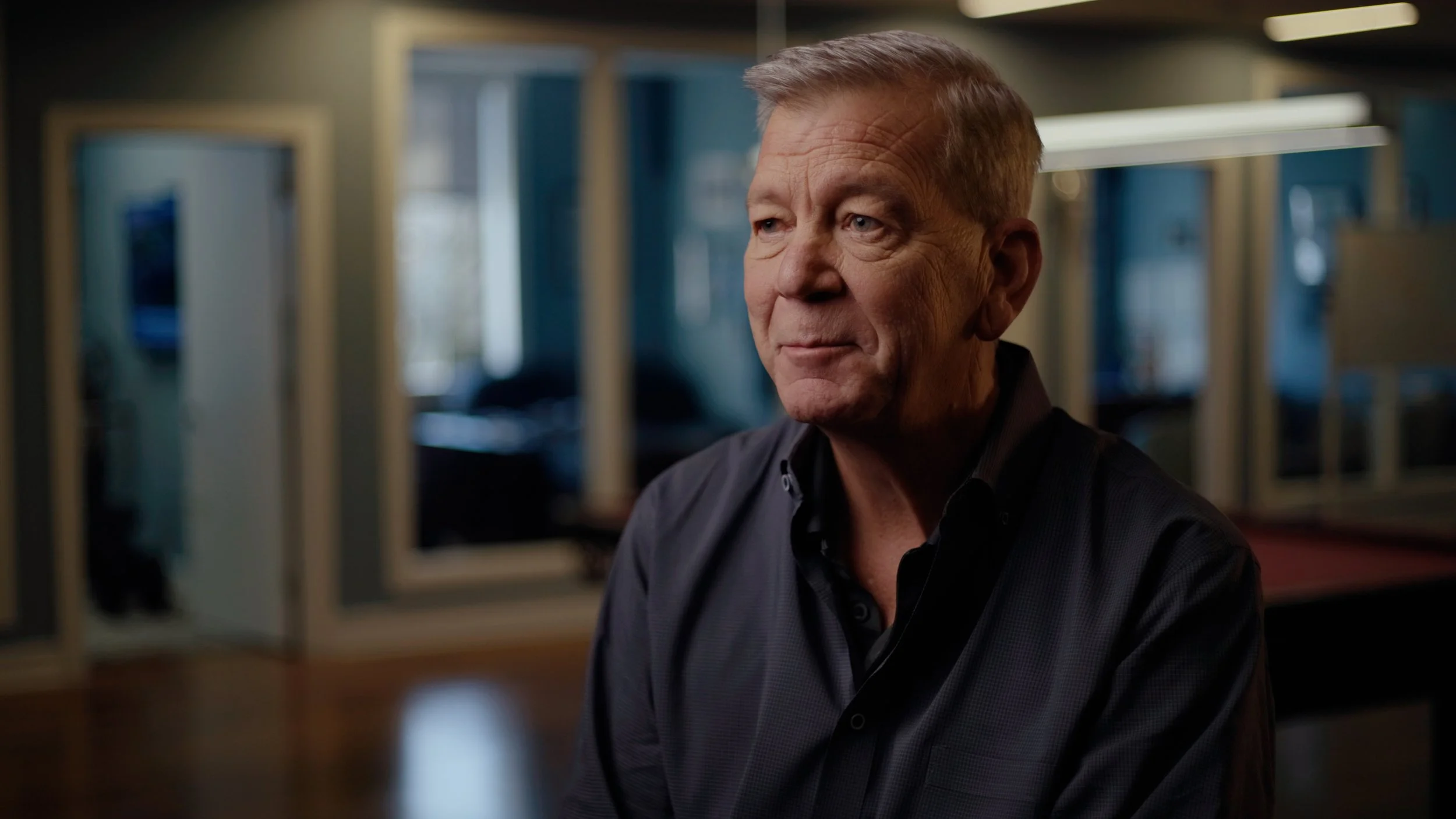 A middle-aged man with short light brown hair, wearing a dark button-up shirt, sitting indoors with a thoughtful expression.