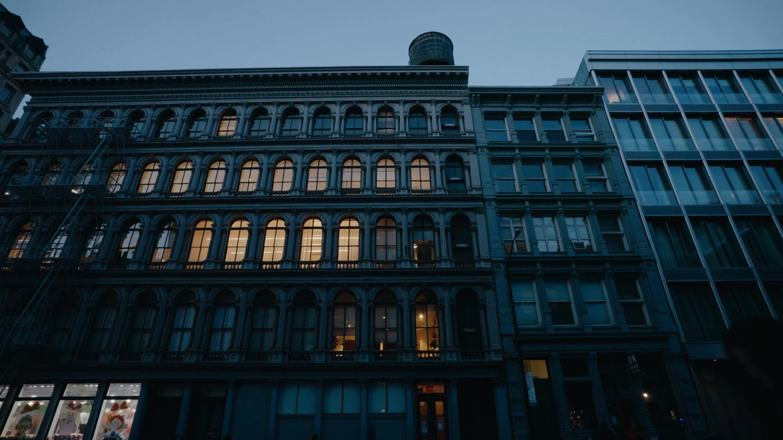 City street view at dusk with multi-story buildings, some illuminated windows, and a cloudy sky.