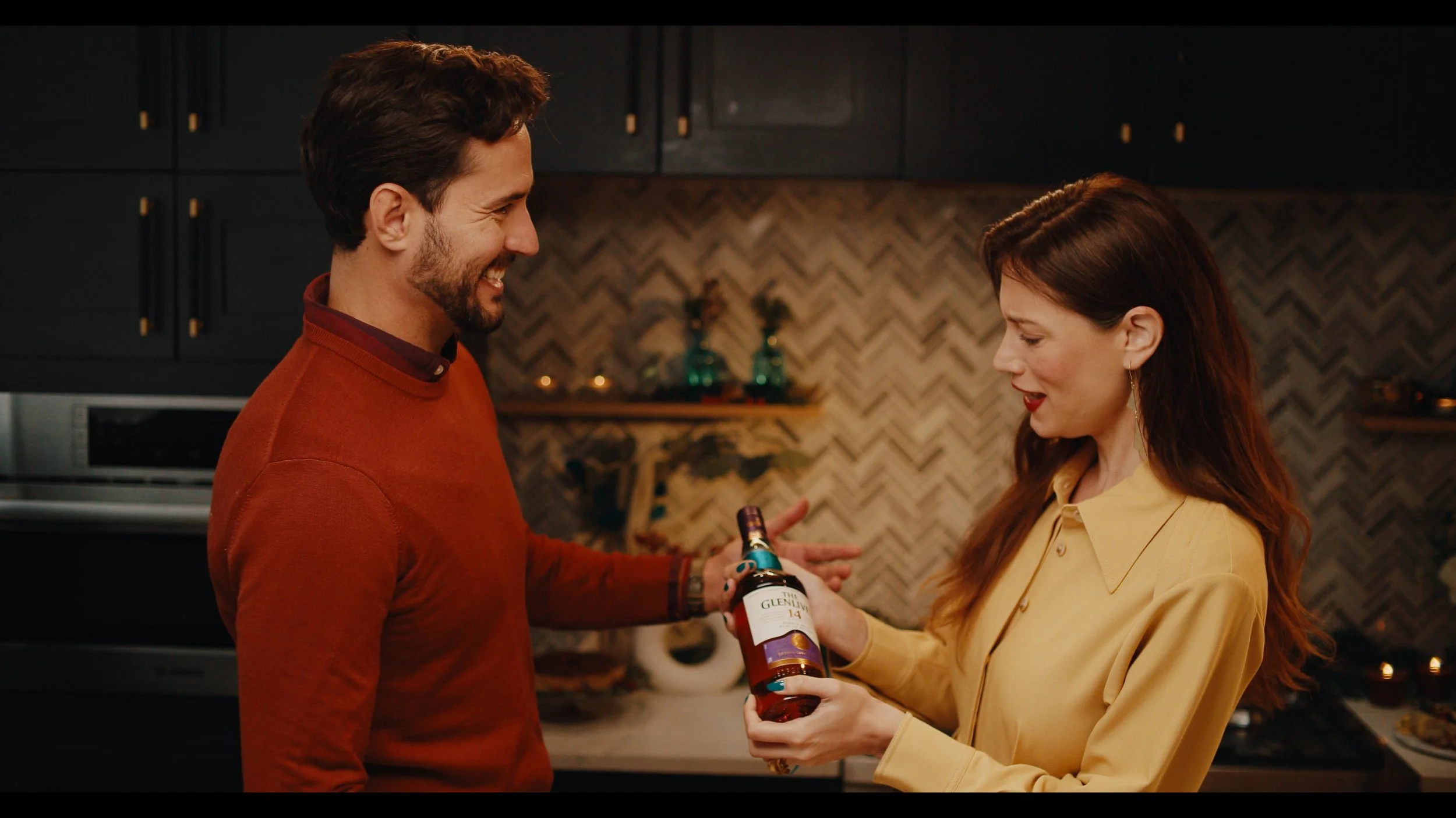 A man with dark hair and a beard smiling at a woman with long red hair as she hands him a bottle of Glenlivet whiskey in a modern kitchen with dark cabinetry and a herringbone backsplash.