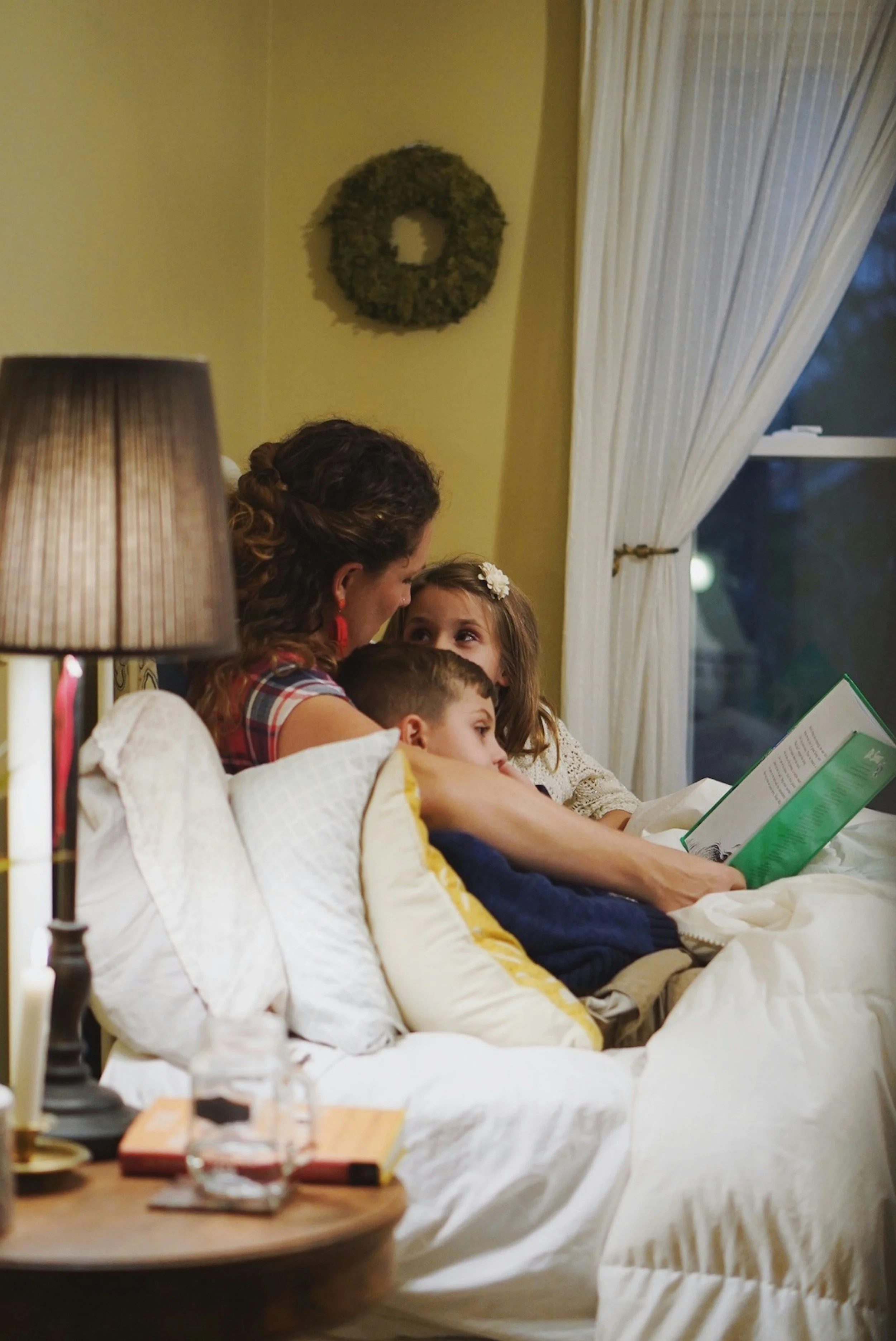 Woman and two children sitting on a bed, reading a book together in a cozy, dimly lit bedroom with a lamp and window with white curtains.