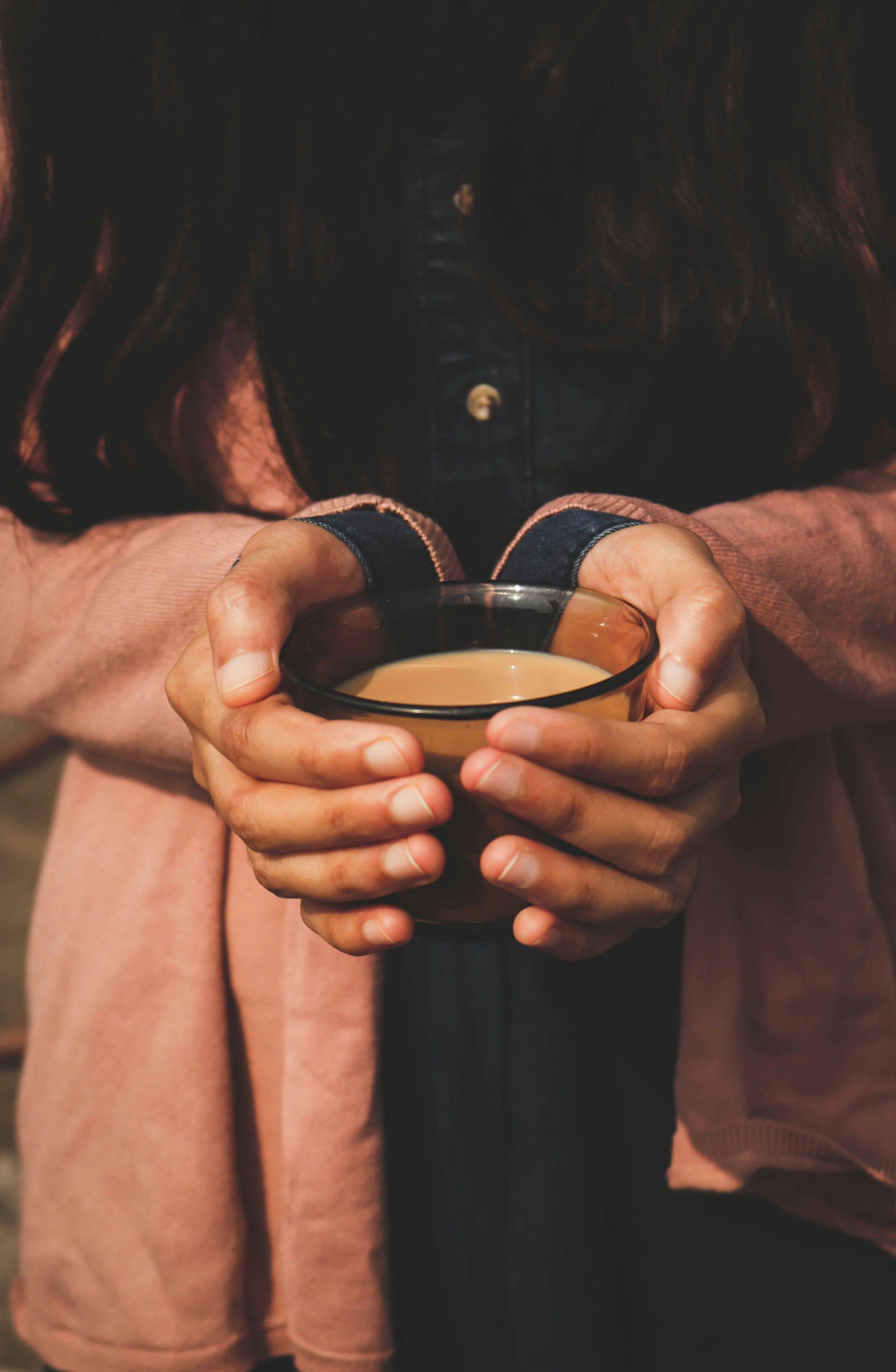 Person holding a glass cup of tea or coffee with both hands, wearing a pinkish-brown coat and black layer underneath.