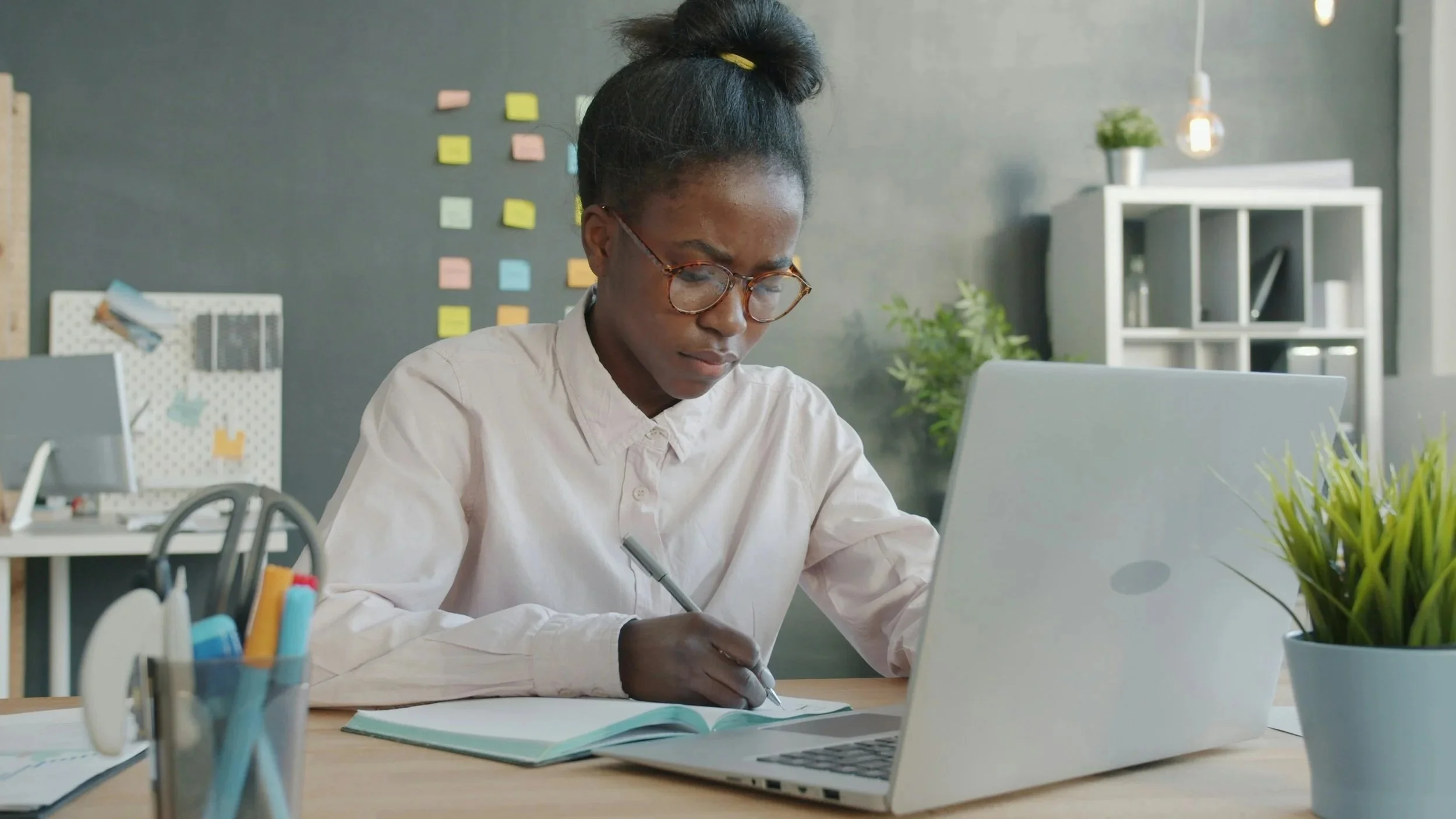 A woman with glasses writing in a notebook at her desk with a laptop, pen holder, and plants in a modern office.