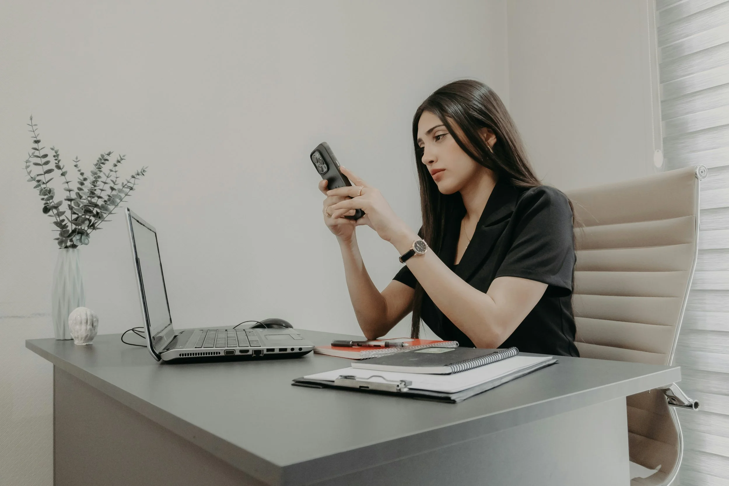 A woman sitting at a desk, looking at her phone, with a laptop, notebooks, and pens on the desk, and a vase of artificial eucalyptus leaves in the background.
