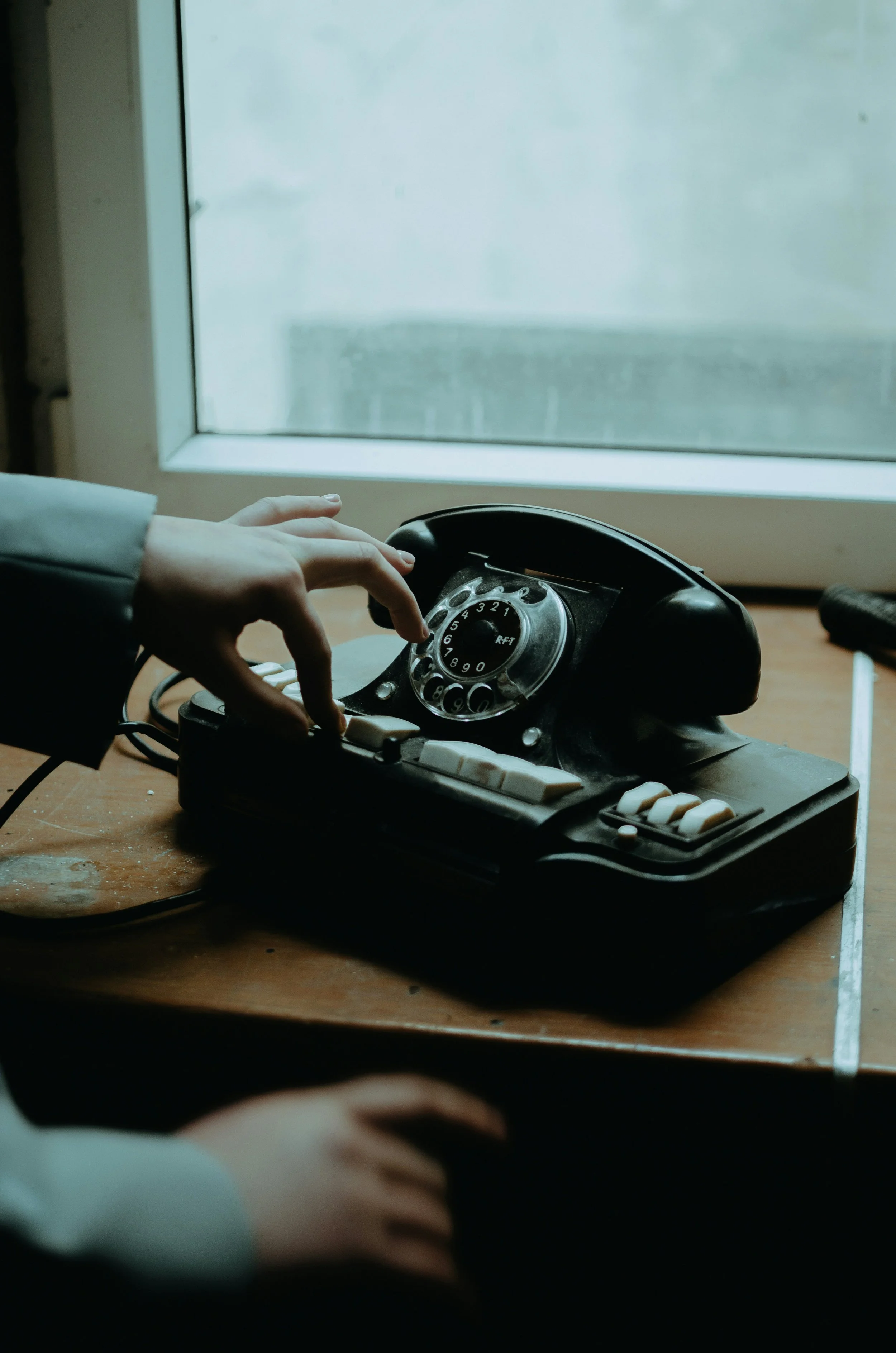 A person operating a vintage black rotary phone on a wooden table near a window.