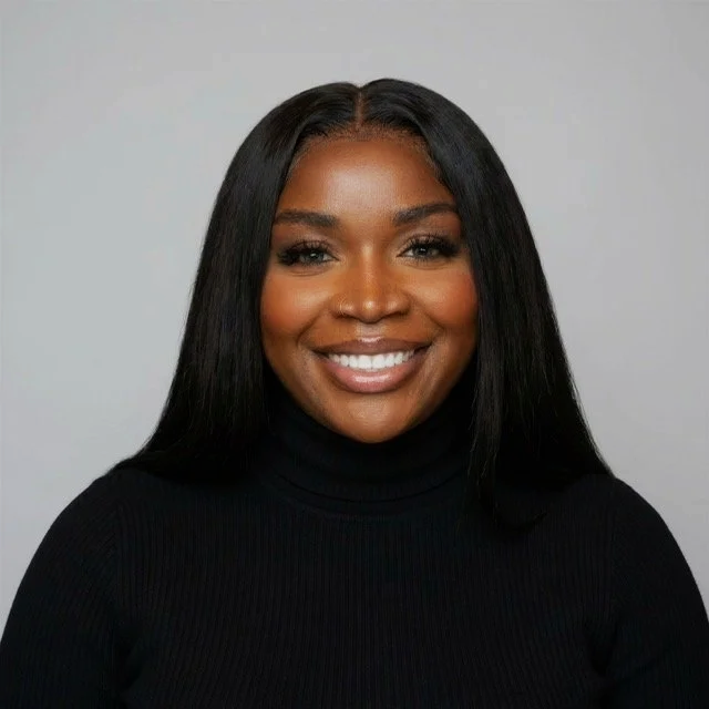 Portrait of a smiling African American woman with straight black hair, wearing a black turtleneck sweater, against a plain light gray background.
