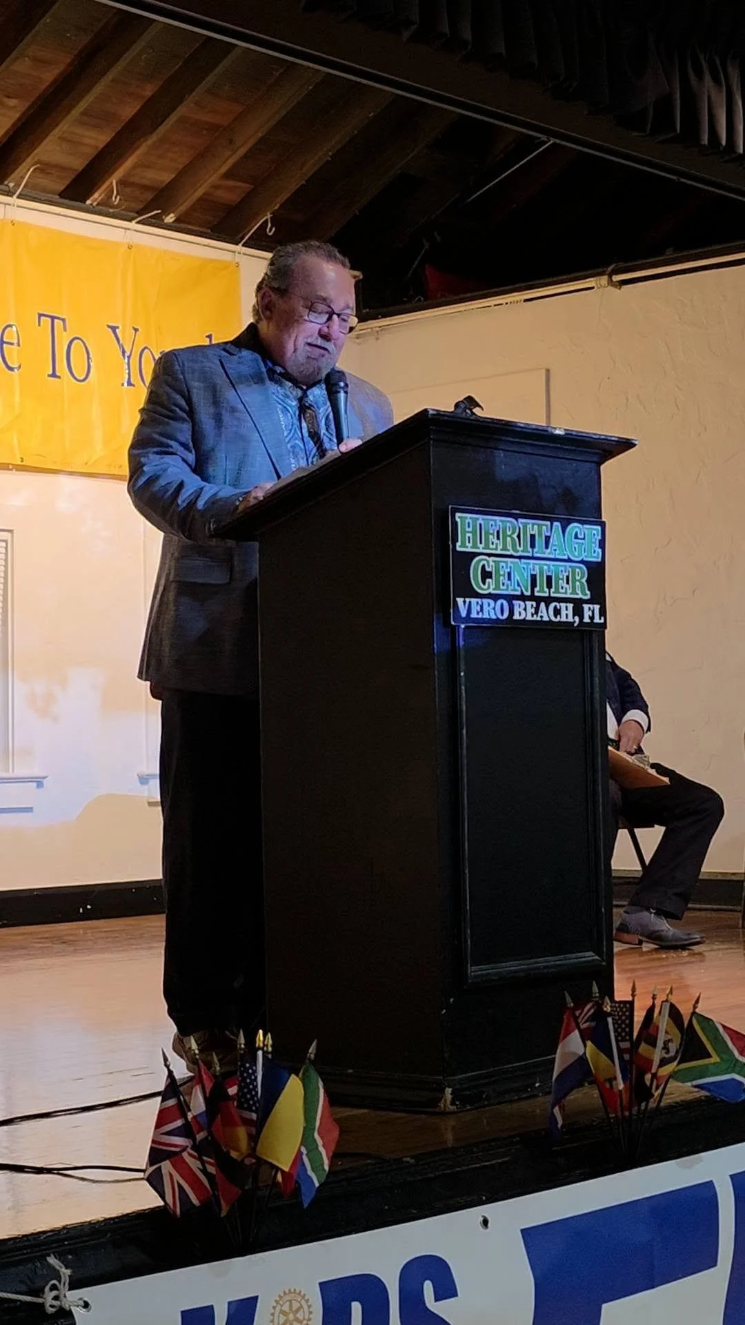 A man in a suit and glasses speaking at a podium with a microphone at the Heritage Center in Vero Beach, Florida. Behind him is a yellow banner with partially visible text, and on the side of the podium is a sign with the center's name. Small flags are displayed at the front of the stage, representing various countries.