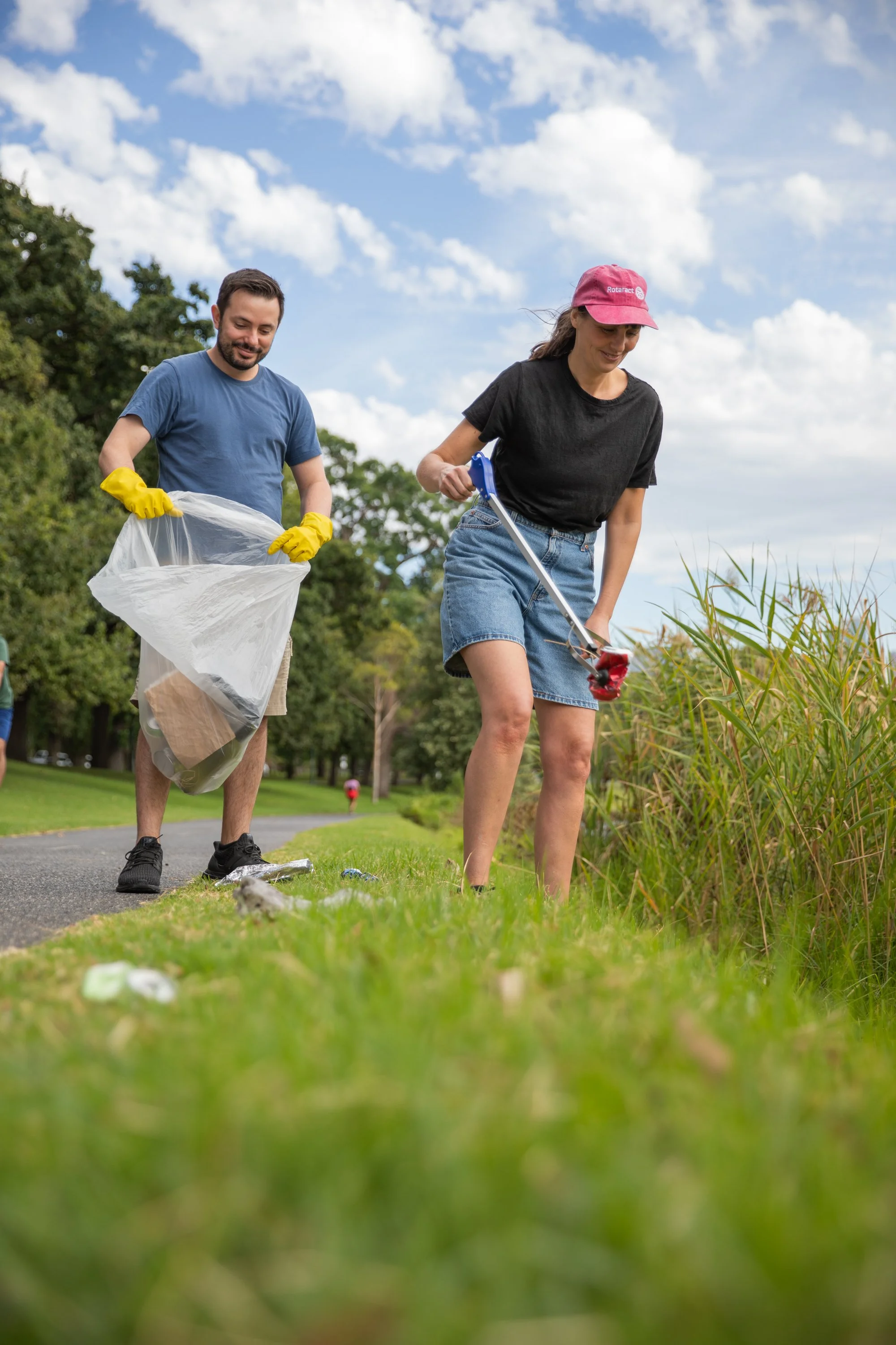 Two volunteers picking up trash along a park sidewalk, with one woman using gardening shears to cut branches and a man holding a trash bag, both wearing gloves and casual clothes.