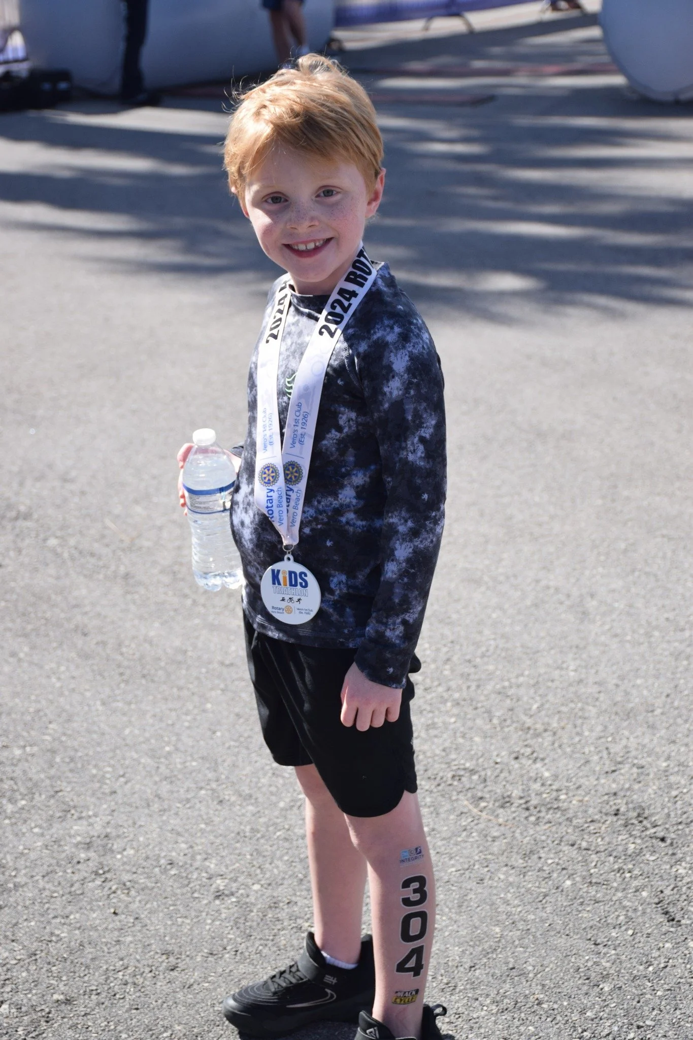 A young boy with red hair, freckles, and a wide smile, standing outdoors on a sunny day. He is holding a water bottle, wearing a race medal around his neck, and has a bib number on his leg. In the background, there are inflatable arches and shadows on the ground.
