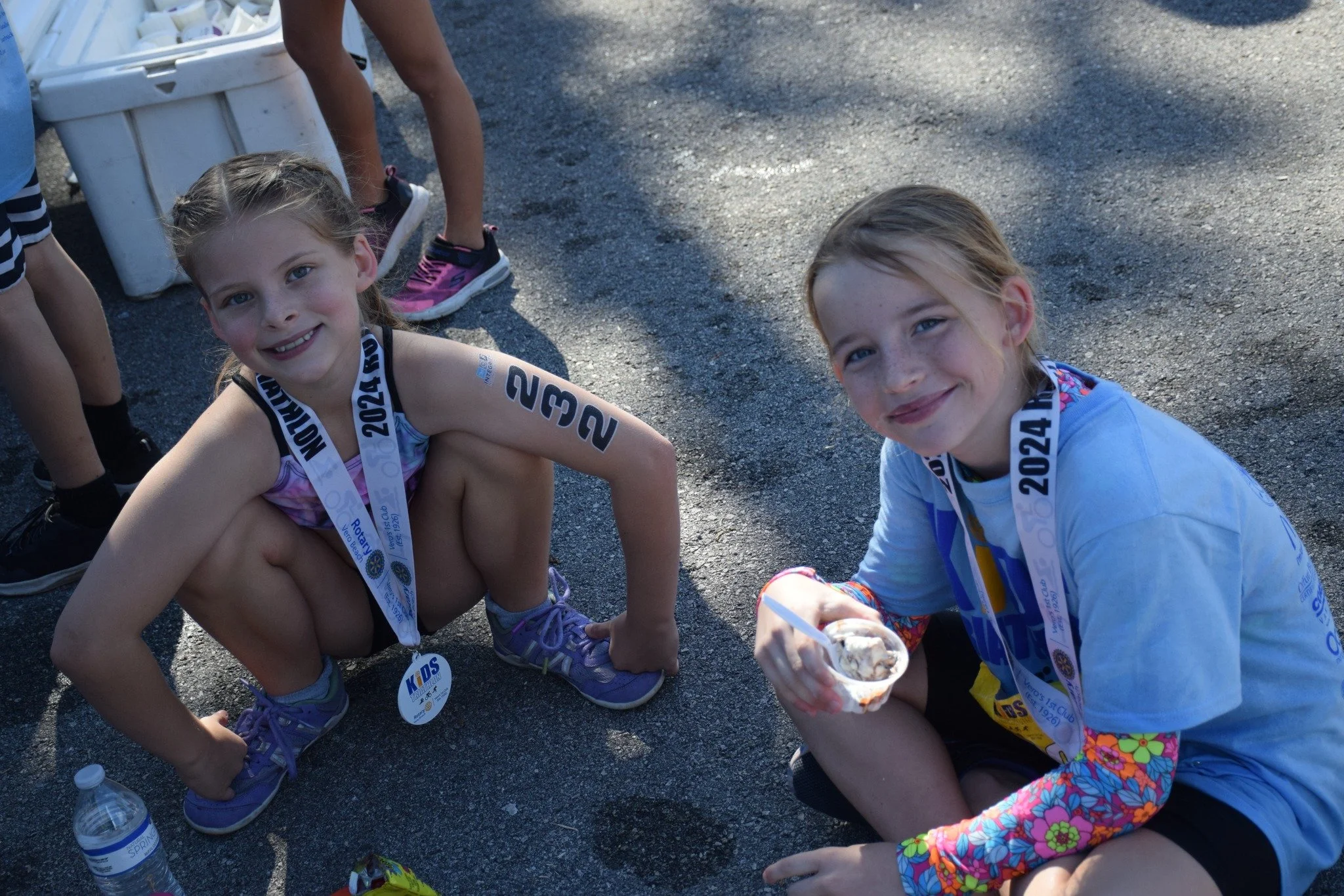 Two young girls sitting on the ground, smiling at the camera. They are wearing medals around their necks, indicating participation in a race or event. One girl is holding a cup of ice cream, and there are water bottles nearby. The background shows other children and gravel ground.
