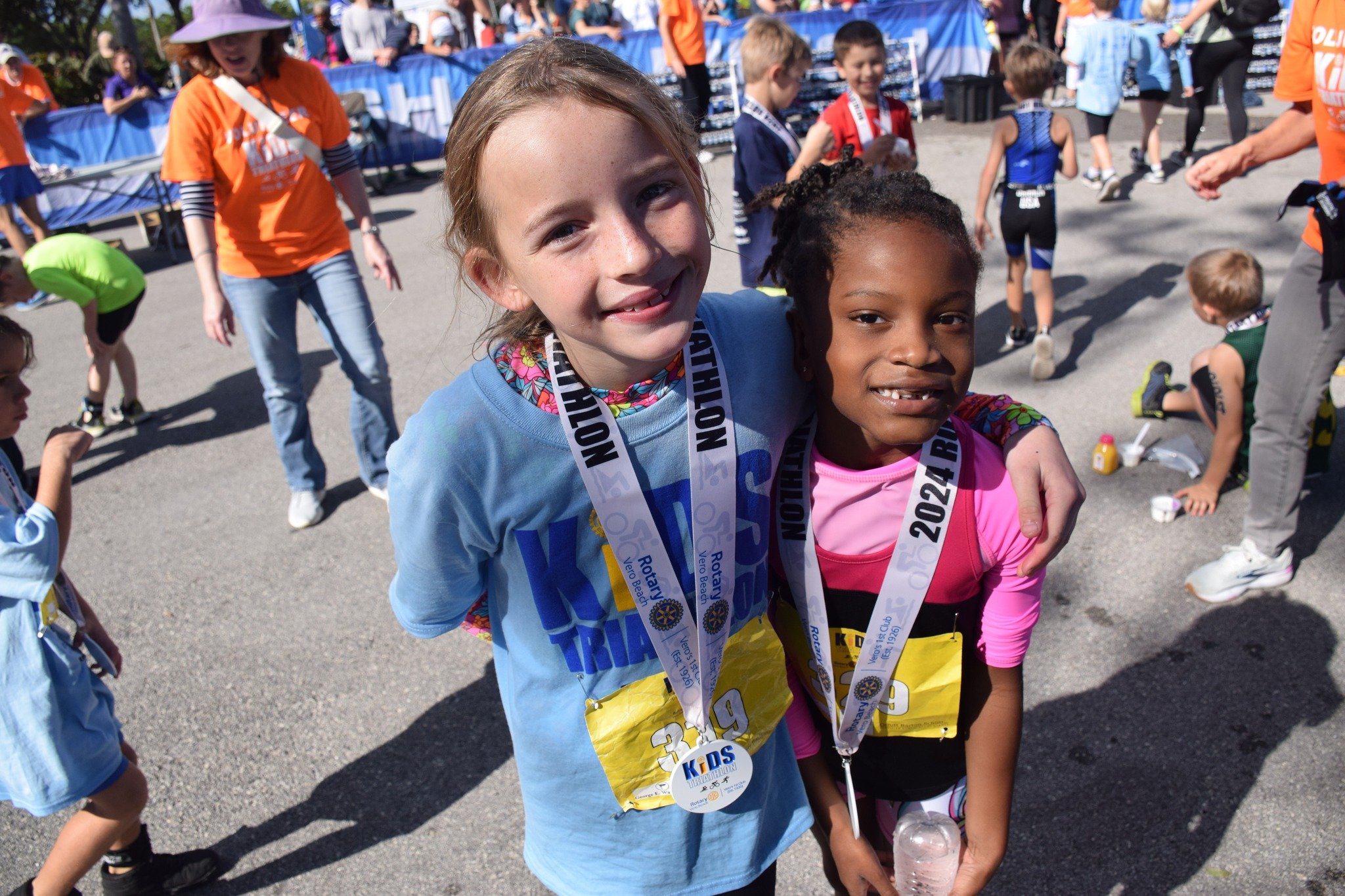 Two young girls wearing race medals and race bibs, smiling and standing close together at a children's race event, with other children and race volunteers in the background.