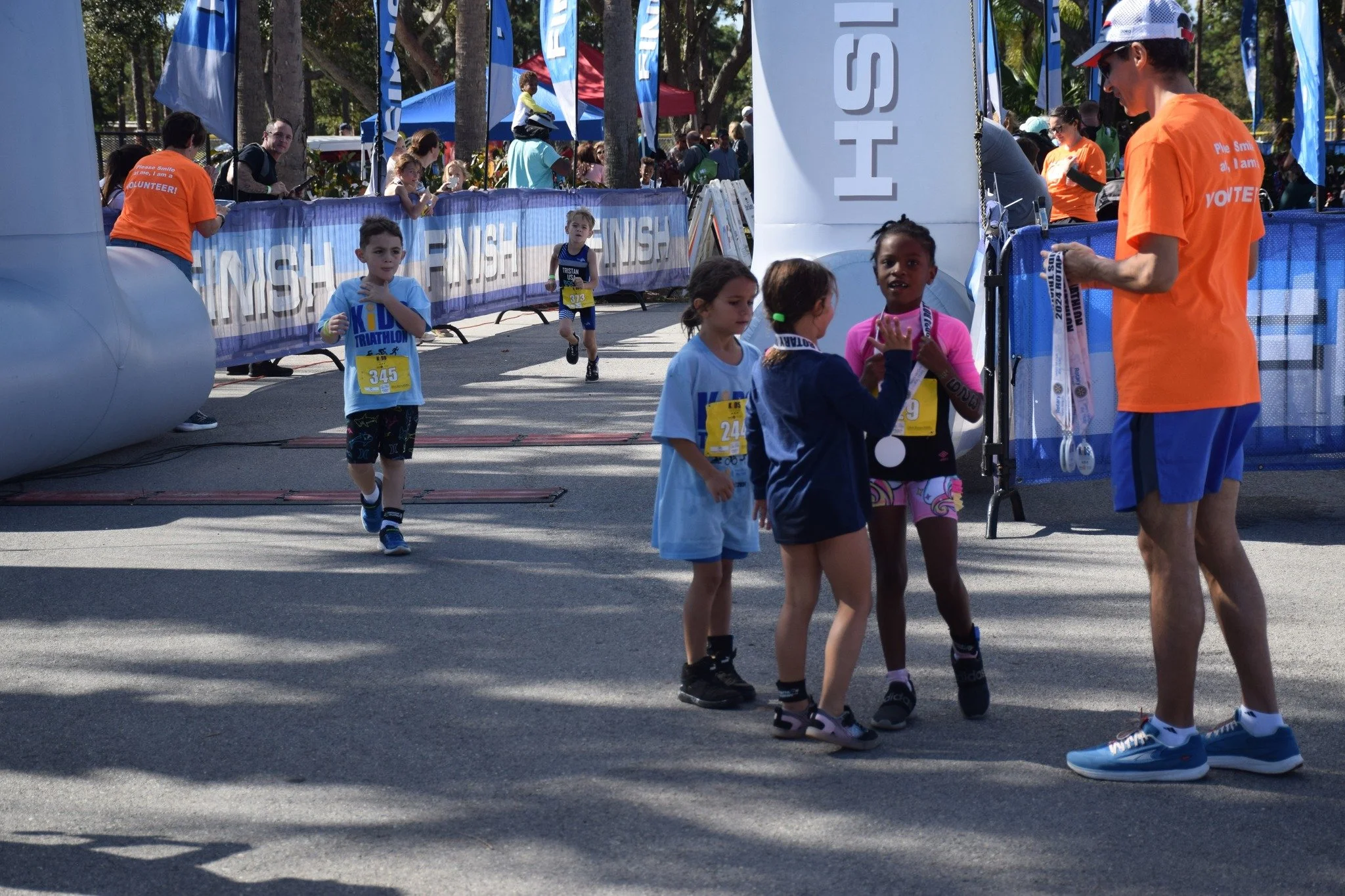 Children and adults participating in a kids' triathlon race. Some children are running towards the finish line, others are standing near the finish area, which is decorated with blue banners and flags. There are spectators and volunteers present, with some wearing orange shirts.