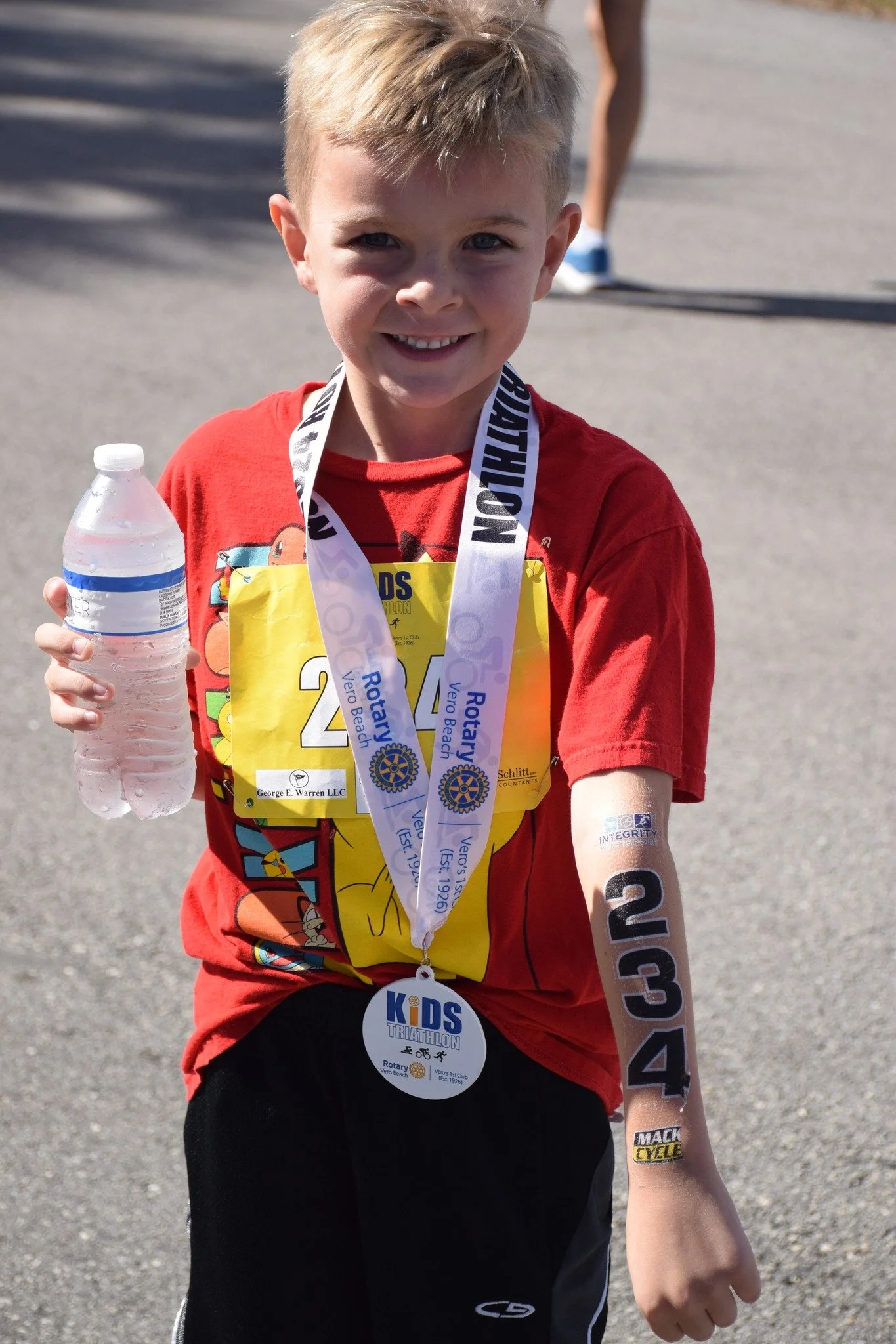 Young boy wearing a red shirt, black shorts, and running medals around his neck, holding a water bottle, smiling at the camera after a race.