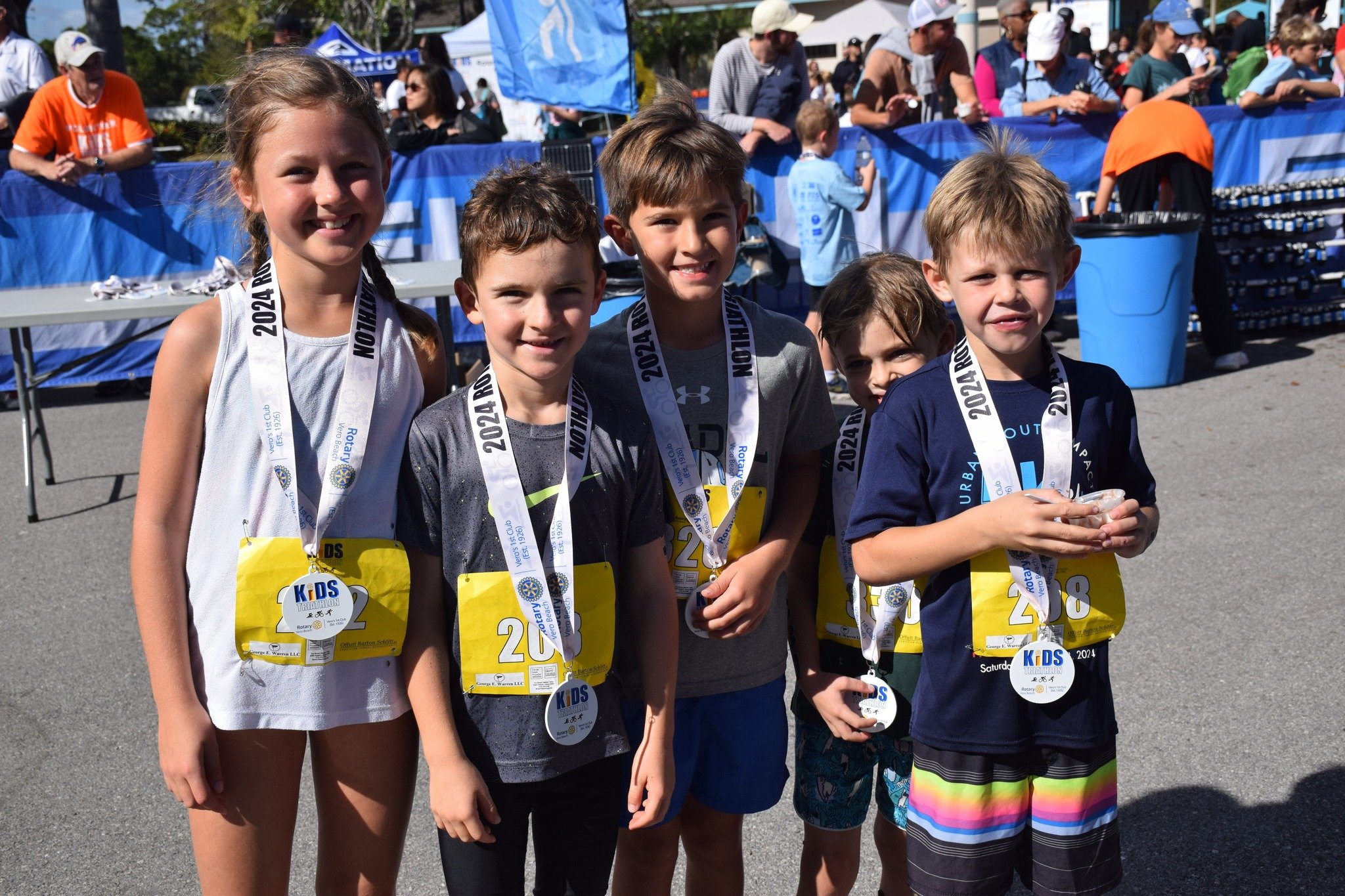 Group of children at a triathlon event, wearing medals and race bibs, standing outdoors with a crowd in the background.