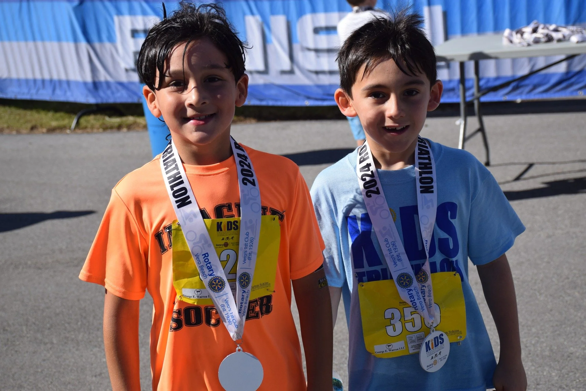 Two young boys standing side by side outdoors, wearing race bibs and medals, smiling at the camera.