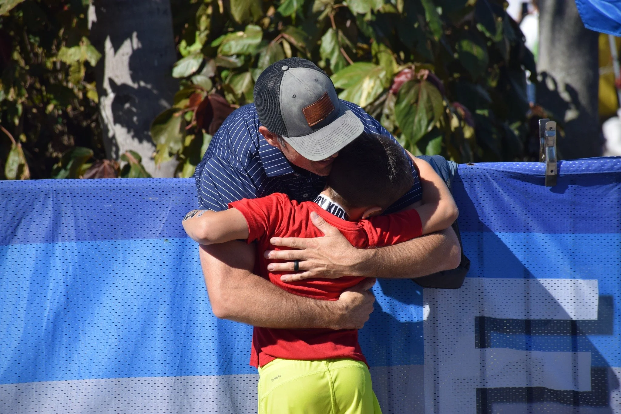 A man in a striped shirt and a black and grey baseball cap hugging a young boy in a red shirt with the yellow shorts, outside near green foliage.