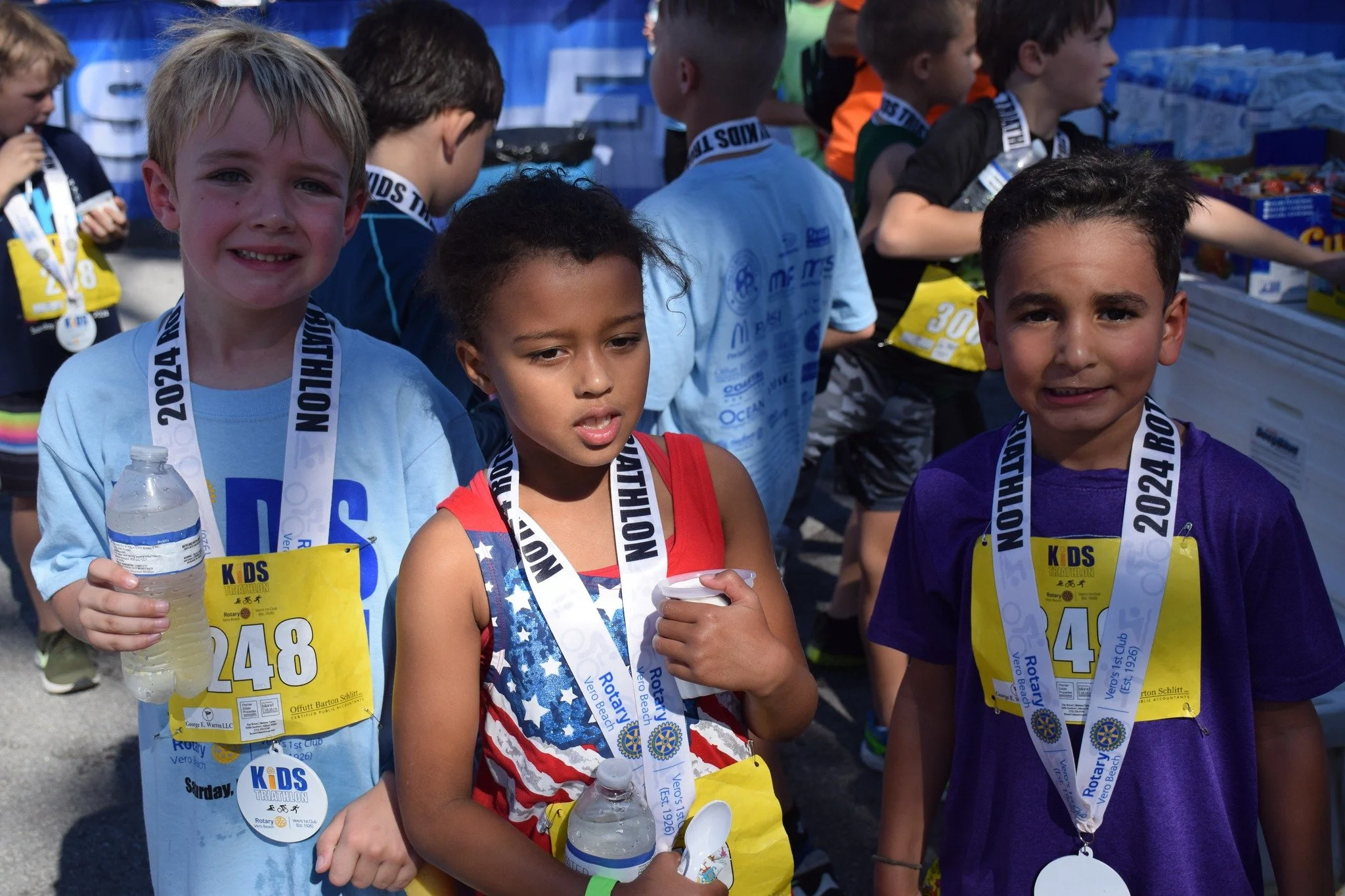 Group of children at a kids' triathlon event, wearing race bibs and medals, some holding water bottles.