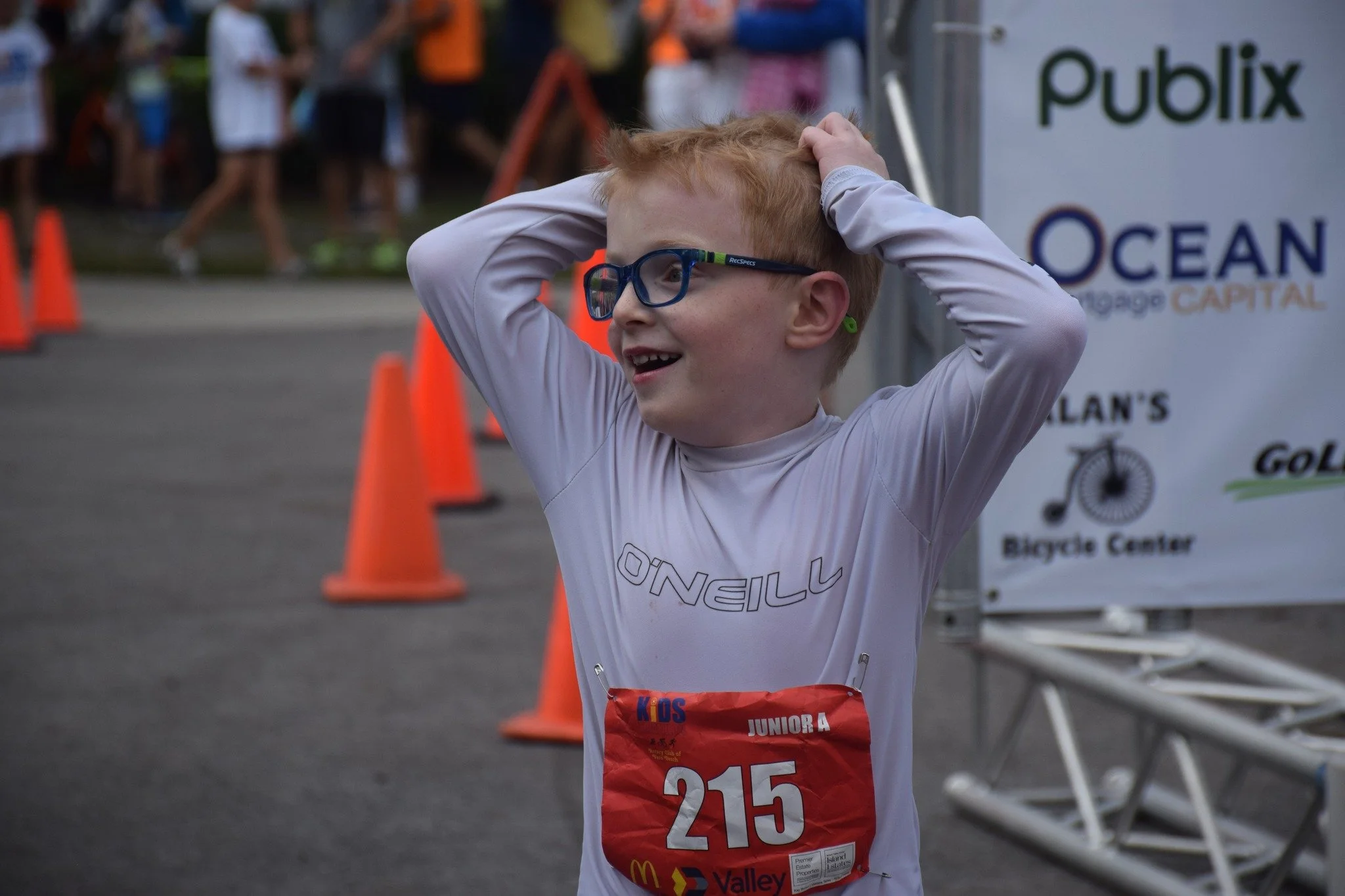 A young boy with red hair, glasses, and a race bib smiling with his hands on his head during a race event.
