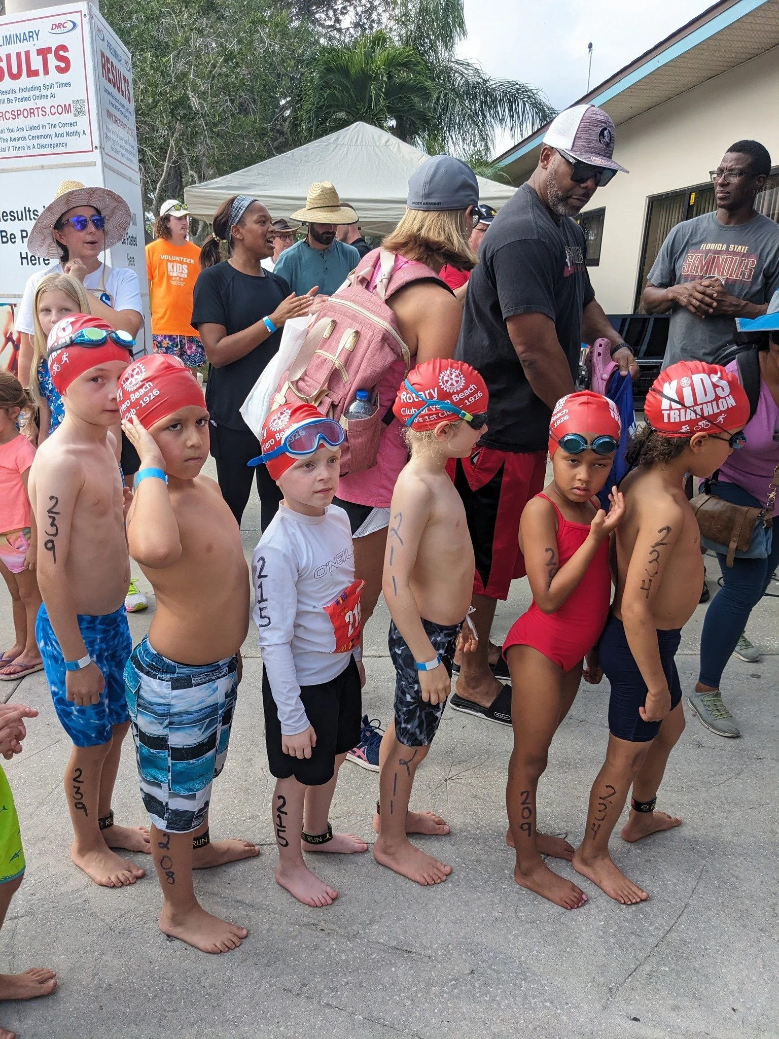 Children in swimwear with handwritten race numbers standing barefoot, preparing for a race outdoors, with adults and a tent in the background.