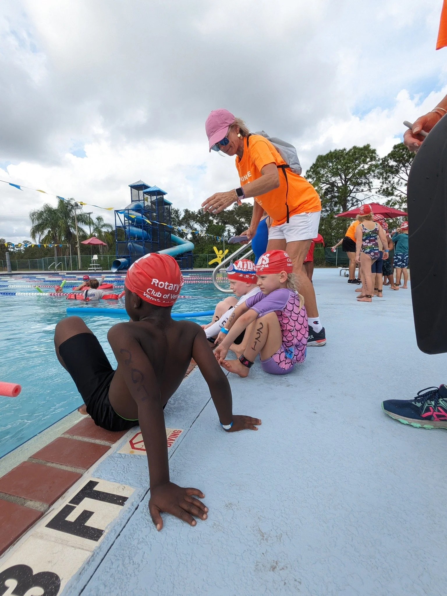 Young swimmers sitting by the edge of a swimming pool, wearing swim caps and bathing suits, while an adult woman in an orange shirt and pink cap helps them prepare to swim. There are people, umbrellas, and a waterslide in the background.