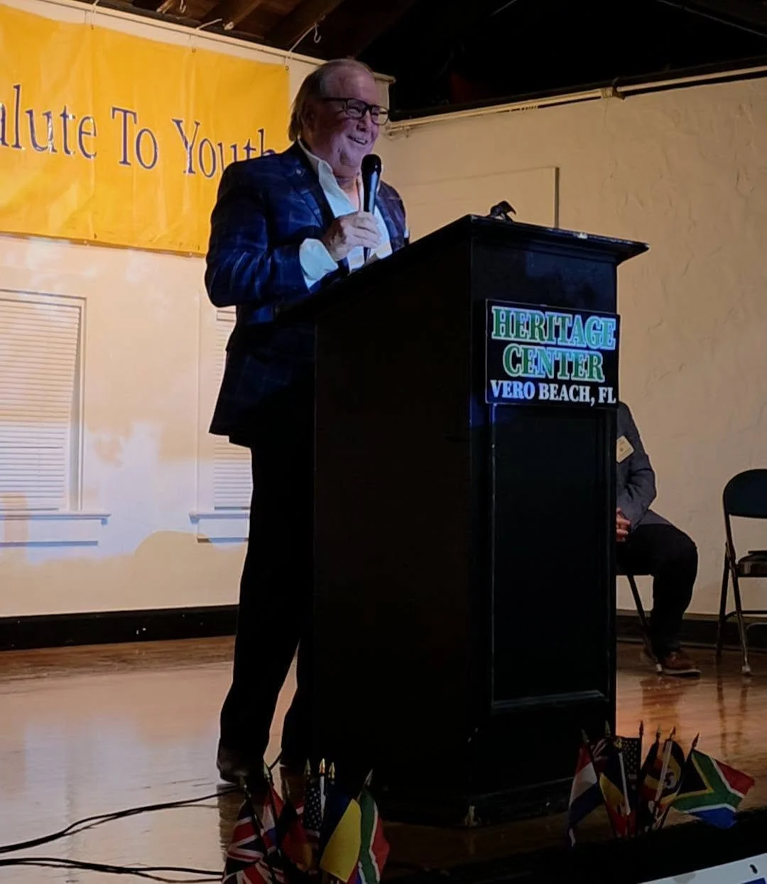 An elderly man wearing glasses and a blue checkered blazer standing behind a black podium with a microphone, speaking at an event at the Heritage Center in Vero Beach, Florida. The background features a yellow banner with partially visible text, and small flags from different countries are placed at the base of the podium.