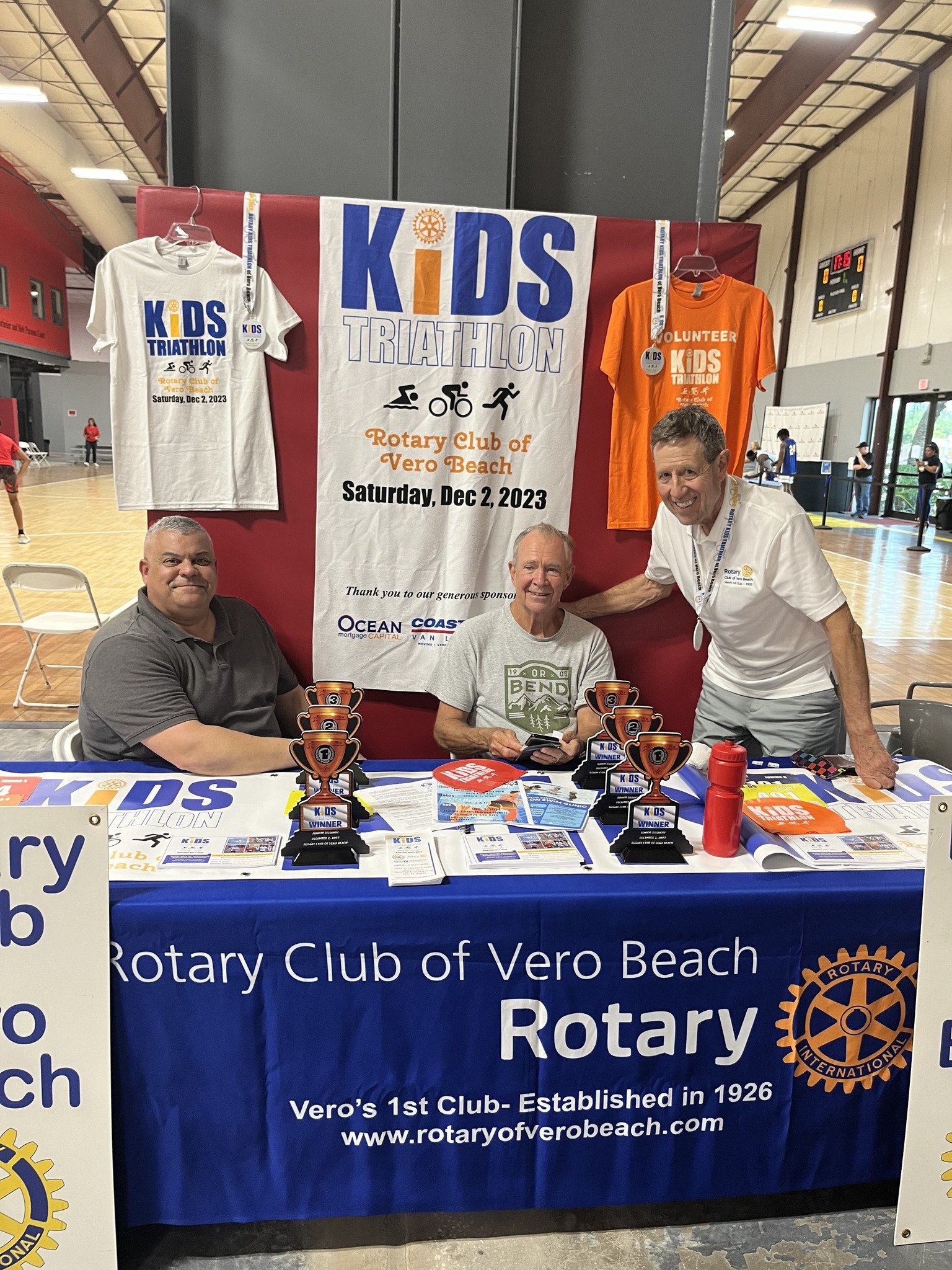 Group of three men sitting in front of a table with trophies and flyers at a Kids Triathlon event organized by the Rotary Club of Vero Beach, held on December 2, 2023, in a gymnasium.